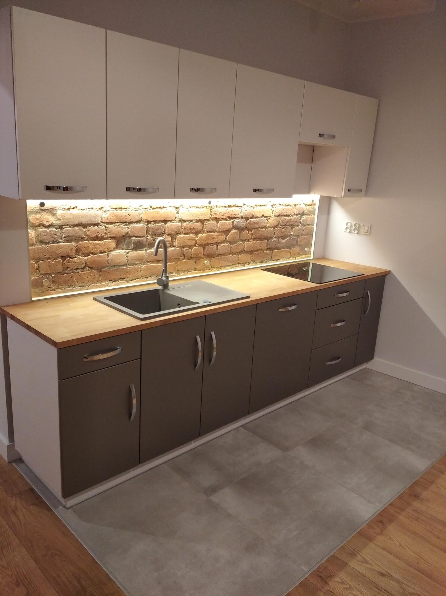 Nowoczesna kuchnia z szarymi szafkami dolnymi i białymi górnymi, drewnianym blatem oraz ceglaną ścianą nad blatem, oświetloną taśmą LED.