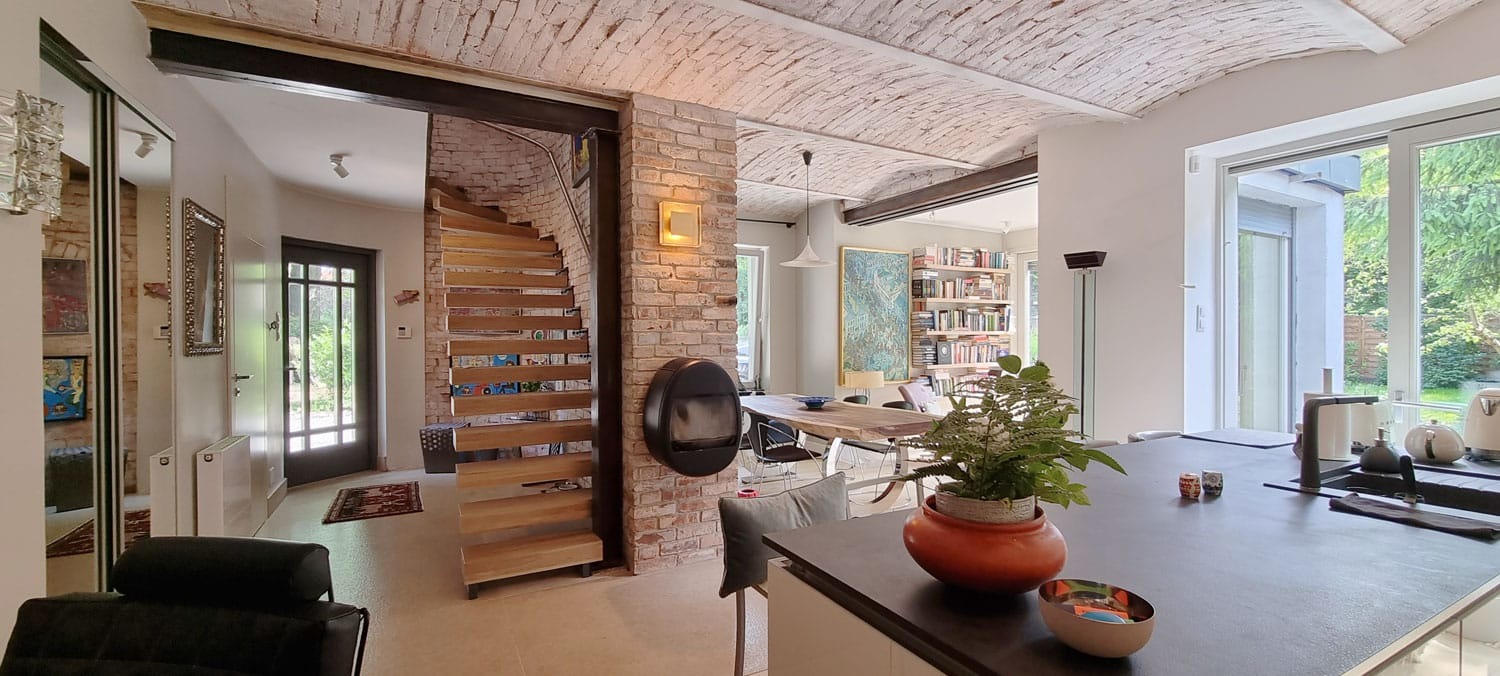 Przestronne wnętrze z ceglanym sklepieniem, drewnianymi schodami i nowoczesnym kominkiem. Widok na otwartą kuchnię i jadalnię, naturalne światło.