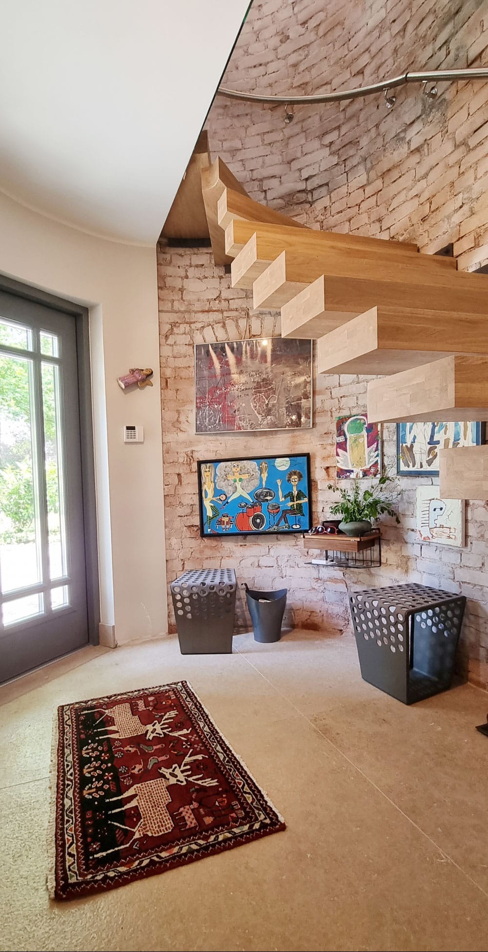 Wnętrze z ceglaną ścianą, drewnianymi schodami i kolorowymi obrazami. Na podłodze wzorzysty dywan, a obok metalowe stołki i kosz. Jasne, przestronne pomieszczenie.