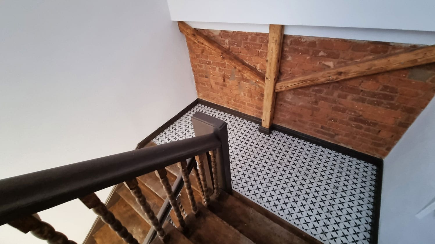 Klatka schodowa po renowacji: białe ściany, wzorzysta podłoga z płytek, drewniane schody i ceglana ściana z drewnianymi belkami. Widok z góry.