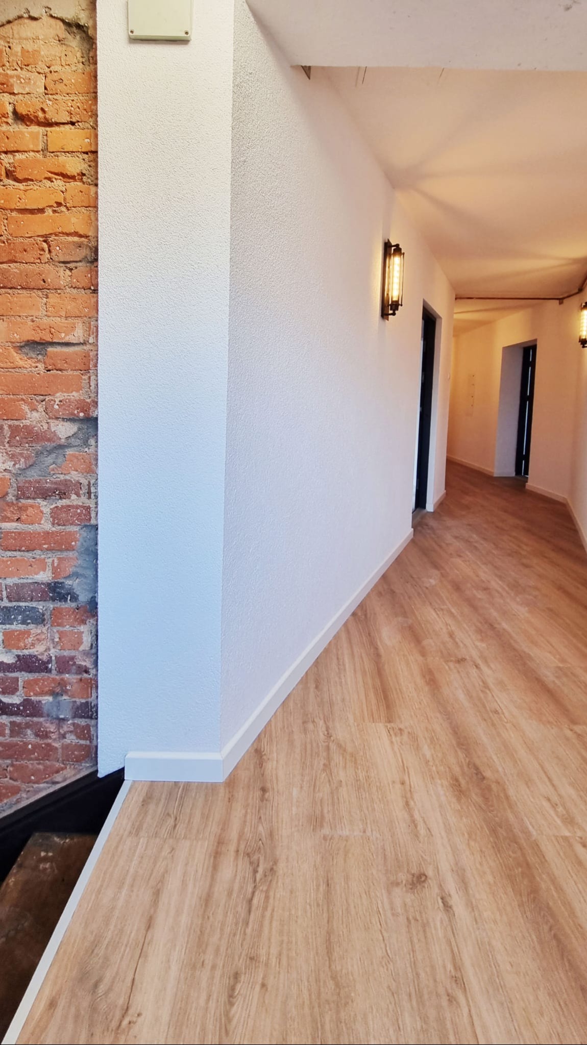 Nowe panele podłogowe w korytarzu, łączące nowoczesne wykończenie ścian z odsłoniętą cegłą. Widoczne oświetlenie ścienne i fragmenty drzwi.