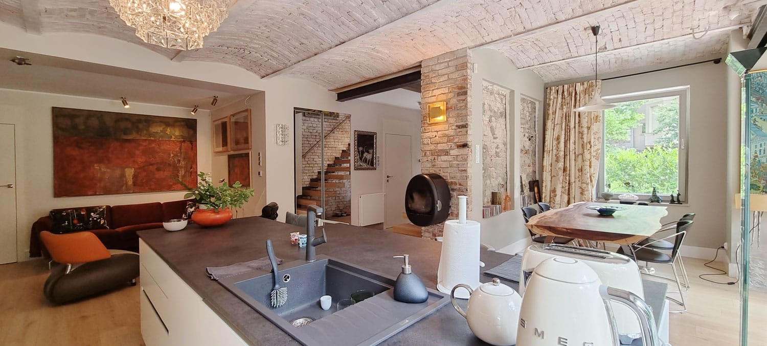 Panoramiczny widok na stylową kuchnię z wyspą, ceglanym sklepieniem i elementami dekoracyjnymi. Widoczny salon z sofą i obrazem.