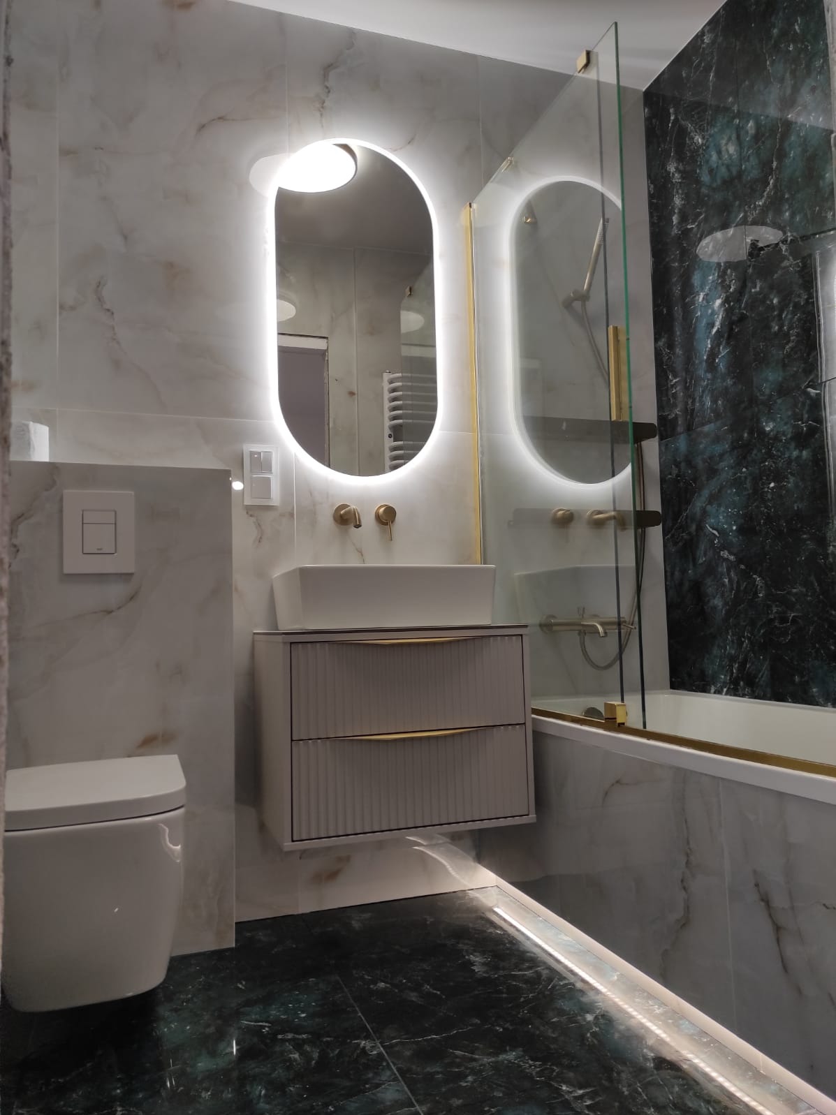 Elegancka łazienka z owalnym lustrem LED, białą umywalką na szafce z frezowanymi frontami i zielono-białą marmurową okładziną. Nowoczesne wykończenie.