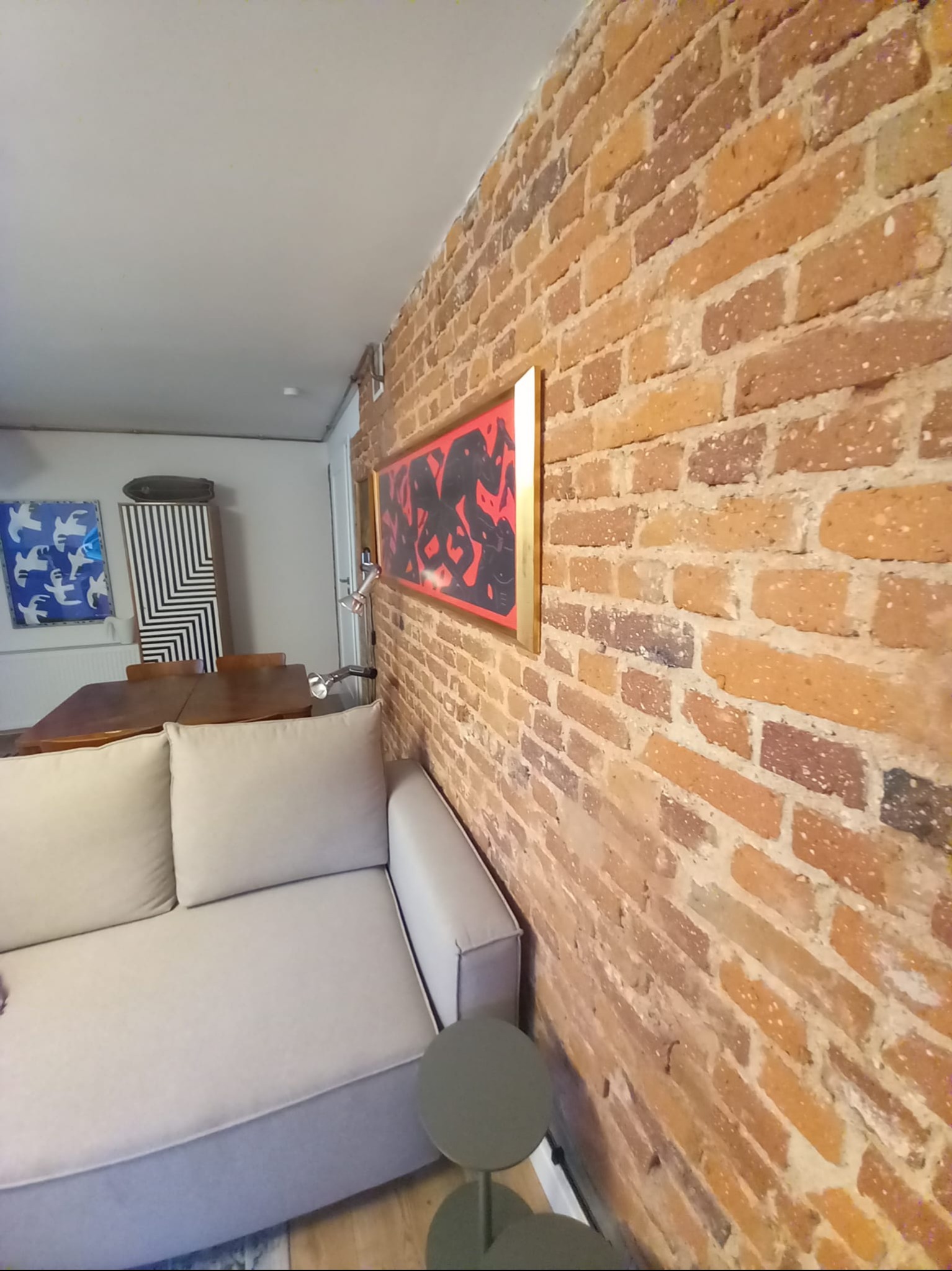 Ceglana ściana w salonie z szarą sofą i nowoczesnymi obrazami. Widoczny fragment stołu i krzesła. Nietypowe oświetlenie podkreśla fakturę cegły.