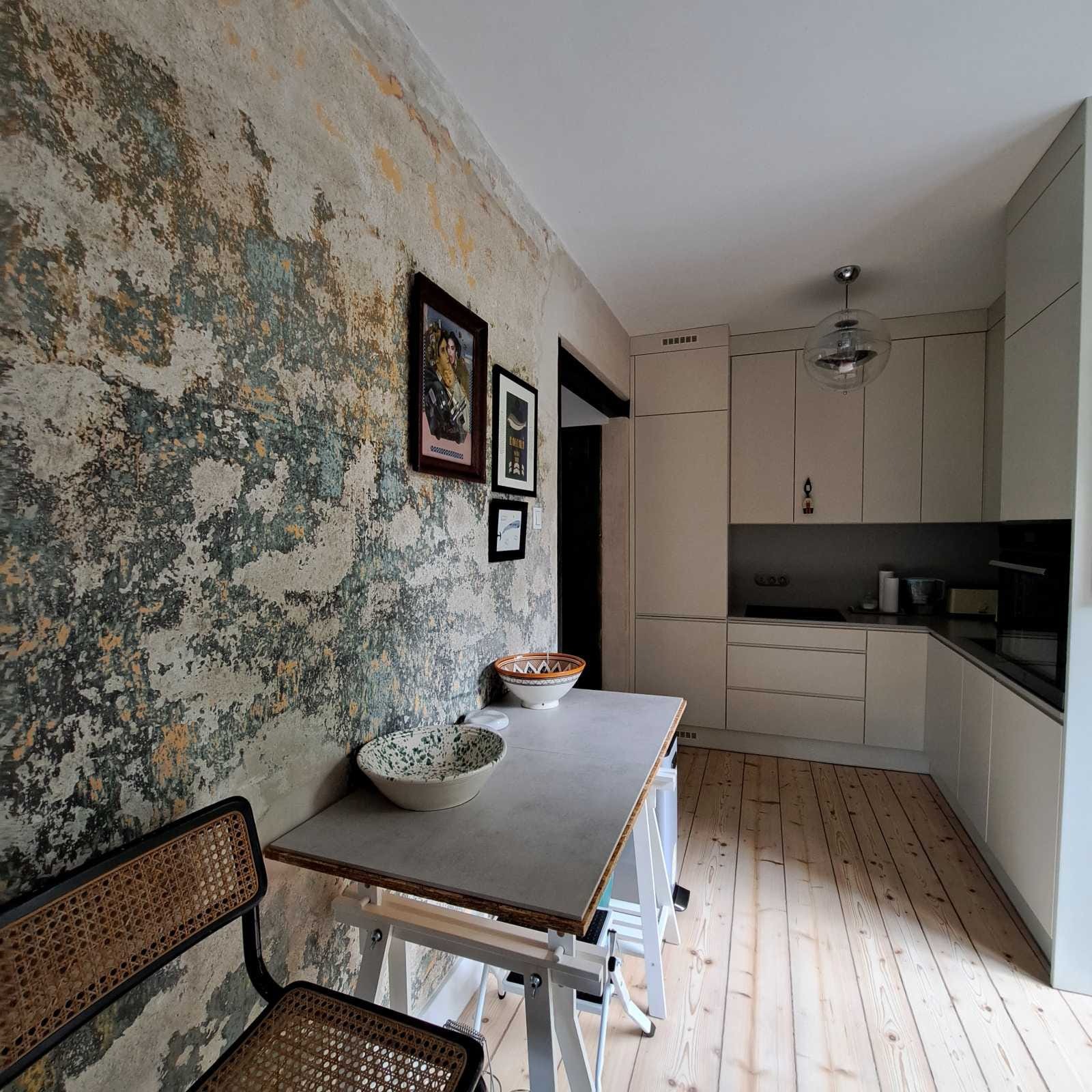 Kuchnia po renowacji: ściana z efektem starego tynku, drewniana podłoga i nowoczesne, jasne meble kuchenne. Na pierwszym planie stół z misami i krzesło.