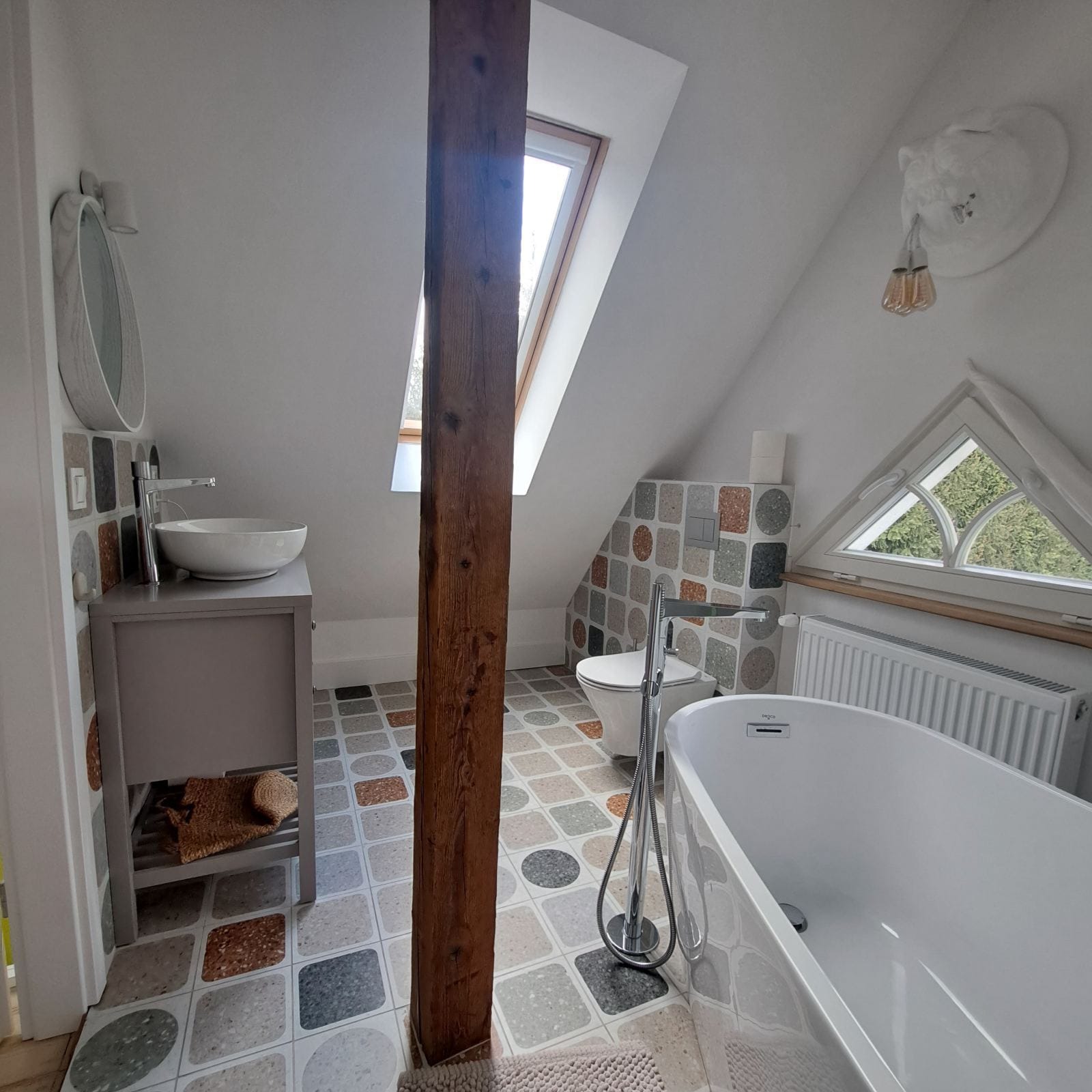 Stylowa łazienka na poddaszu z drewnianą belką, mozaikową podłogą i ścianą, umywalką na szafce i wolnostojącą wanną. Nietypowe okno w kształcie trójkąta.