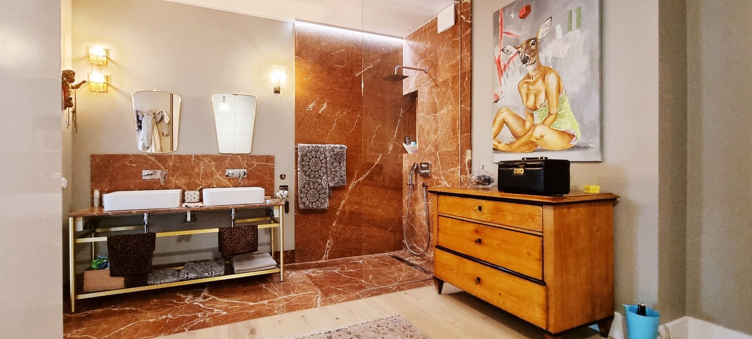 Stylowa łazienka z kabiną prysznicową wyłożoną marmurem, umywalką na złotym stelażu i komodą z obrazem. Nietypowe oświetlenie i dodatki.