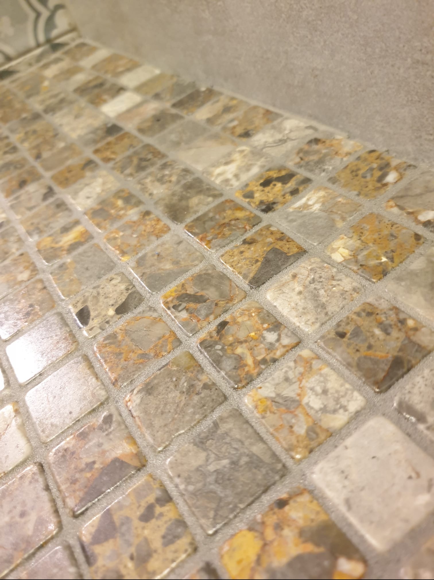 Mozaika z małych, kwadratowych płytek w odcieniach beżu, szarości i brązu, ułożona na powierzchni blatu. Widoczna fuga w jasnym odcieniu. Ujęcie z bliska, pod kątem.