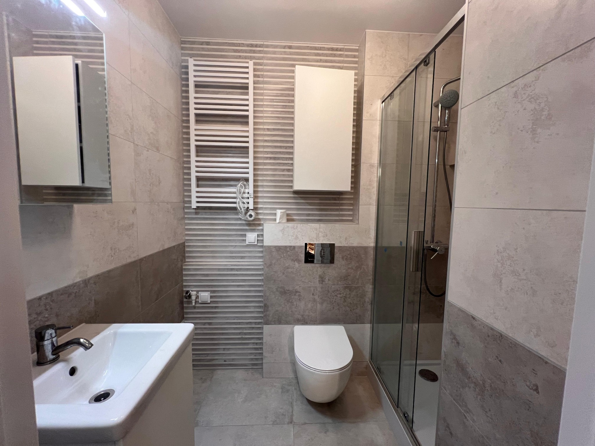 Wykończona łazienka w szaro-białej kolorystyce z kabiną prysznicową, umywalką z szafką, toaletą i grzejnikiem drabinkowym. Widoczne płytki w dwóch odcieniach szarości.