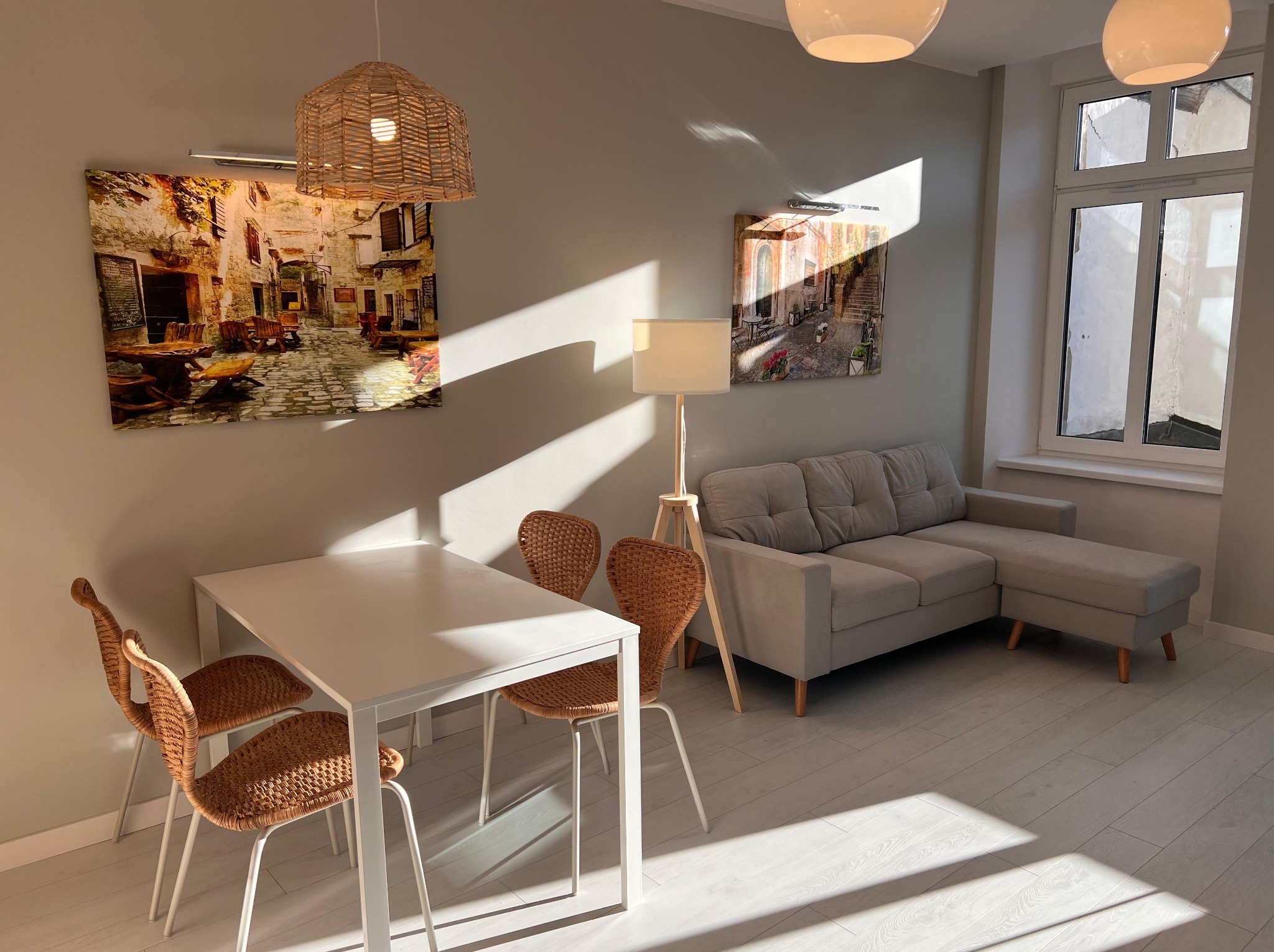Jasne wnętrze z białym stołem, plecionymi krzesłami, szarą sofą i obrazami z motywami miejskimi. Promienie słoneczne wpadają przez okno, oświetlając podłogę.