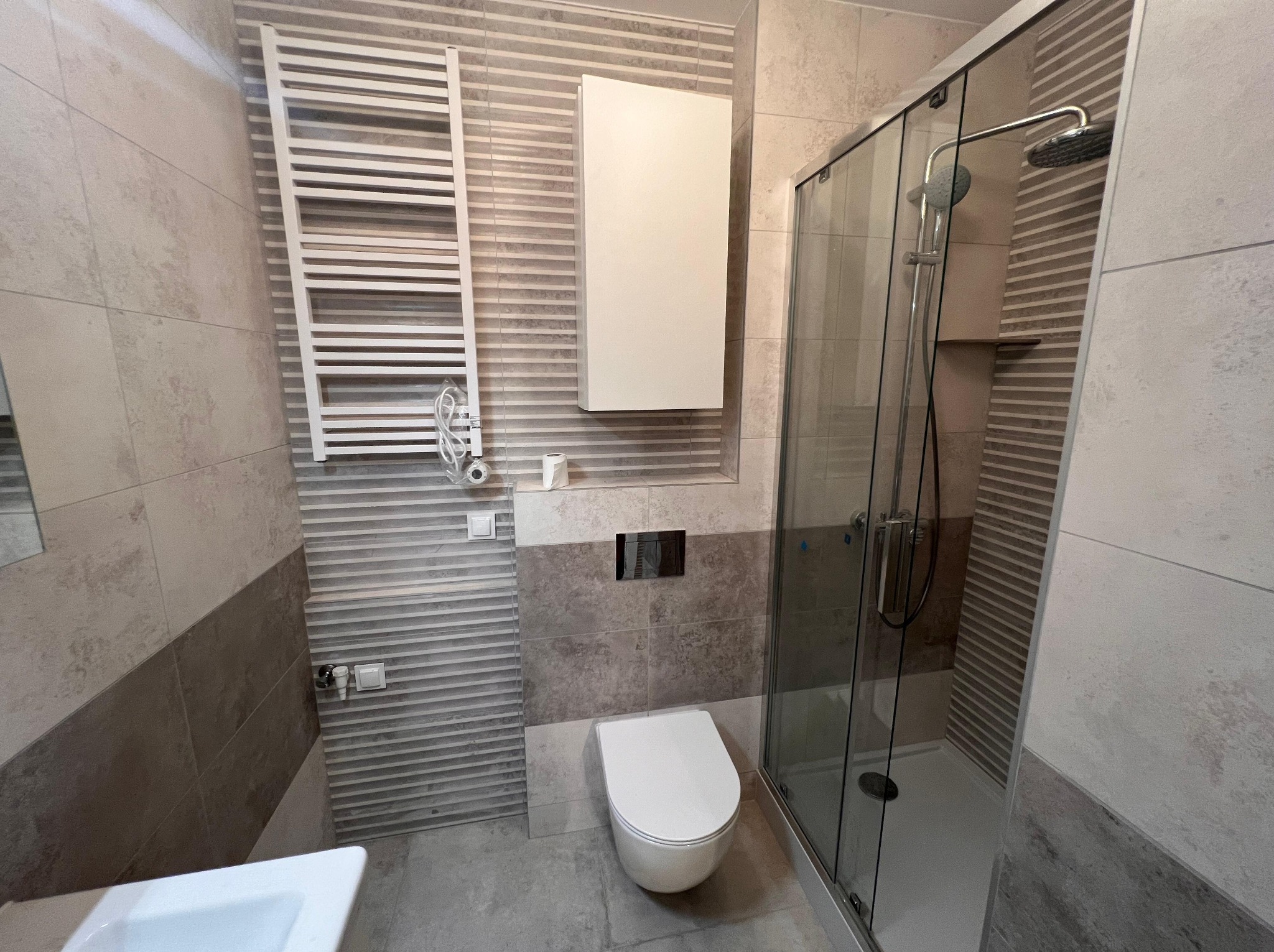 Nowoczesna łazienka po remoncie: kabina prysznicowa ze szklanymi drzwiami, podwieszana toaleta, grzejnik drabinkowy i szafka wisząca na tle płytek w odcieniach szarości i beżu.