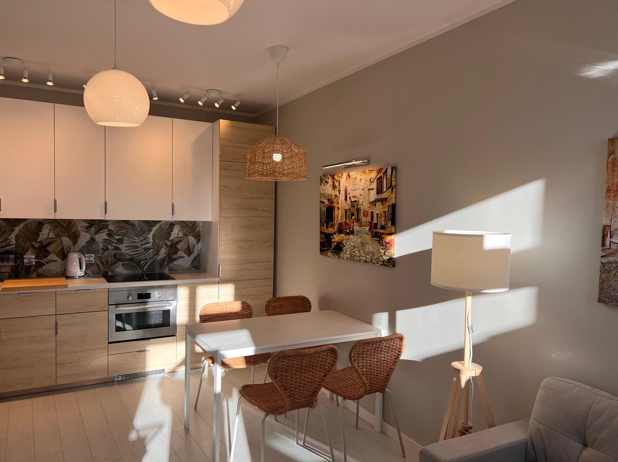 Jasne wnętrze: kuchnia z drewnianymi szafkami i wzorzystą ścianą, biały stół z plecionymi krzesłami, lampa podłogowa i obraz na ścianie w ciepłym świetle słonecznym.