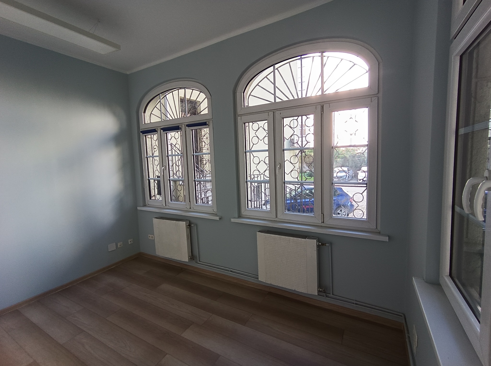 Pomalowane na jasny odcień szarości wnętrze z drewnianą podłogą i dwoma oknami łukowymi z dekoracyjnymi kratami. Grzejniki pod oknami. Widok na zewnątrz.