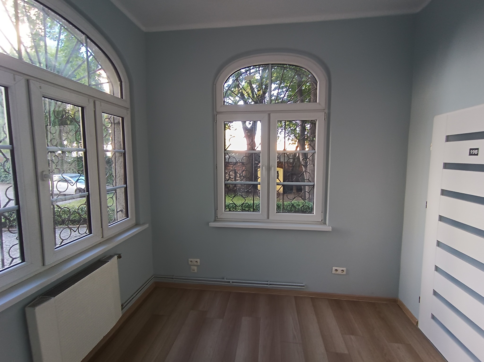 Pomalowane na pastelowo ściany pokoju z dwoma oknami o łukowatych ramach i grzejnikiem pod jednym z nich. Na podłodze panele, widoczna biała, nowoczesna stolarka drzwiowa.