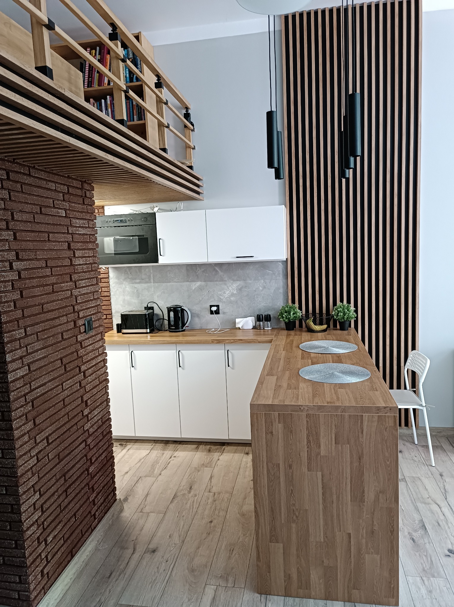 Nowoczesna kuchnia z ceglanym filarem, drewnianym blatem i lamelami na ścianie. Widoczne białe szafki, antresola z książkami i designerskie lampy.