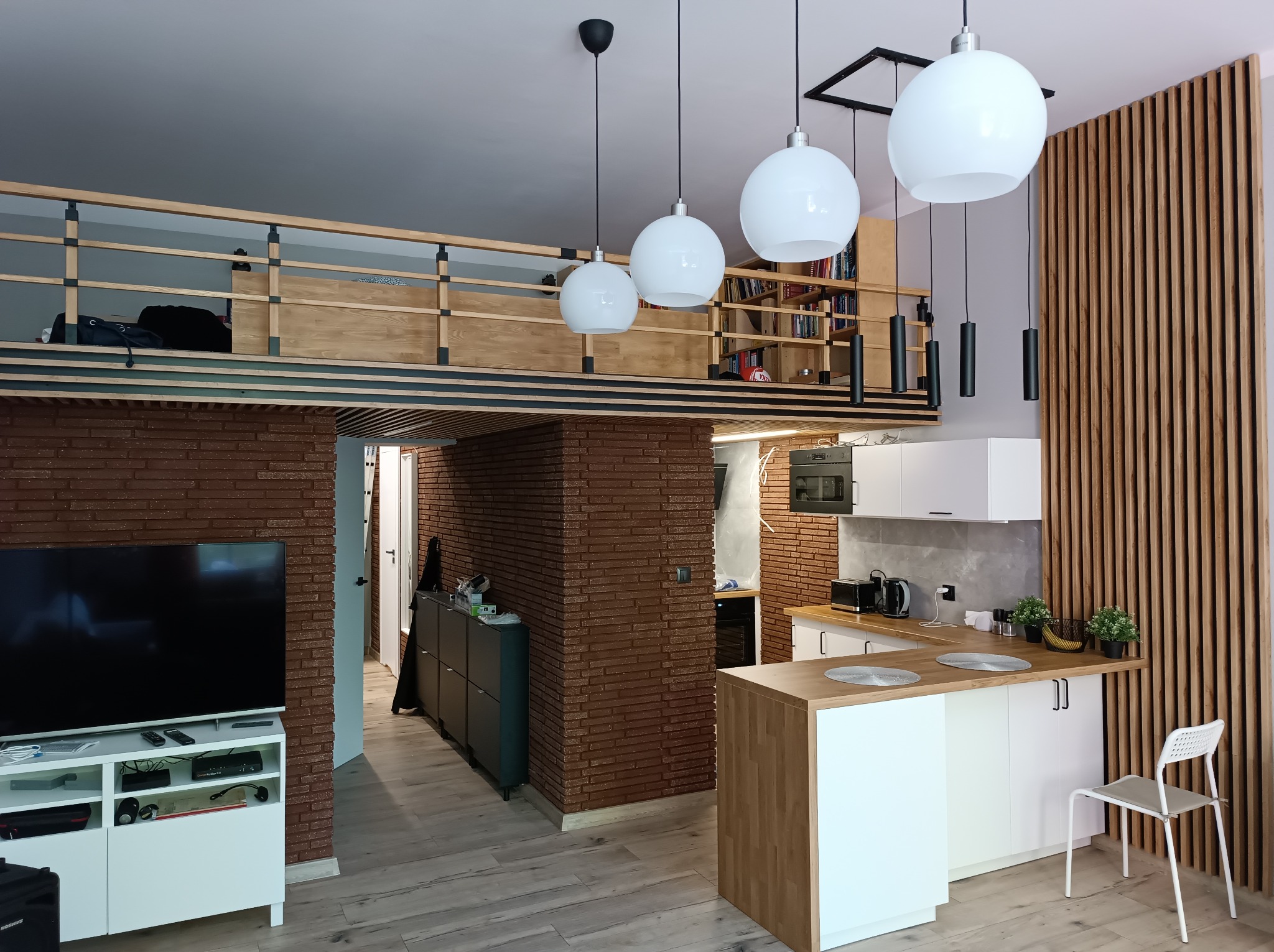 Nowoczesne wnętrze z antresolą, ceglaną ścianą i drewnianymi lamelami. Biała kuchnia z wyspą i okrągłymi lampami. Widoczny telewizor i regał z książkami.
