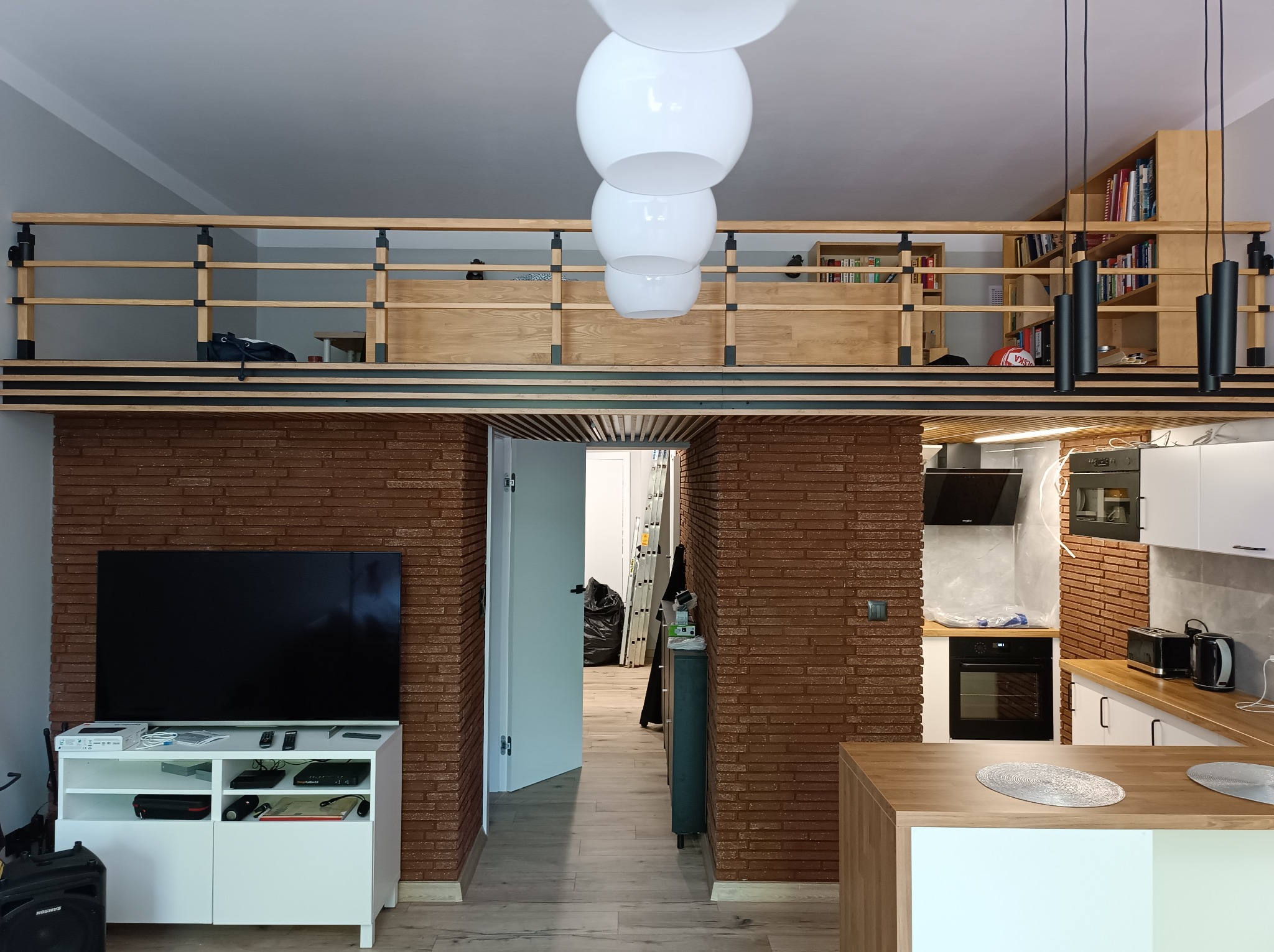 Nowoczesne wnętrze z antresolą i ceglaną ścianą. Widoczny salon z telewizorem, kuchnia z drewnianym blatem i oświetlenie. Całość utrzymana w jasnej kolorystyce.