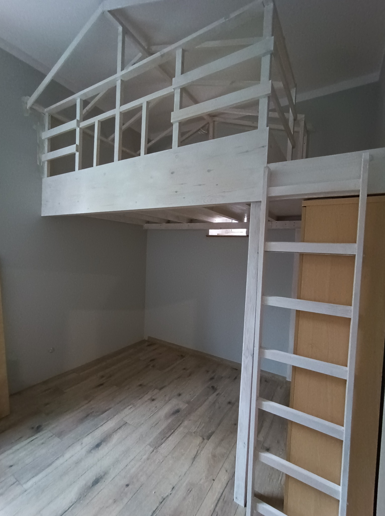 Pokój z drewnianym łóżkiem na antresoli, pomalowanym na biało, z drabinką i szafą wbudowaną pod spodem. Jasne panele na podłodze, szare ściany.