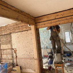Rznijze praca w drewnie - Mężczyzna na drabinie podczas prac remontowych, widoczne odsłonięte belki stropowe, fragment ściany z cegły i instalacje, używany odkurzacz przemysłowy.