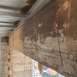 Rznijze praca w drewnie - Drewniana belka stropowa z widocznymi śladami obróbki i częściowo zachowaną starą powłoką, podtrzymywana przez metalową konstrukcję sufitu podwieszanego, w tle fragment ściany z bali.