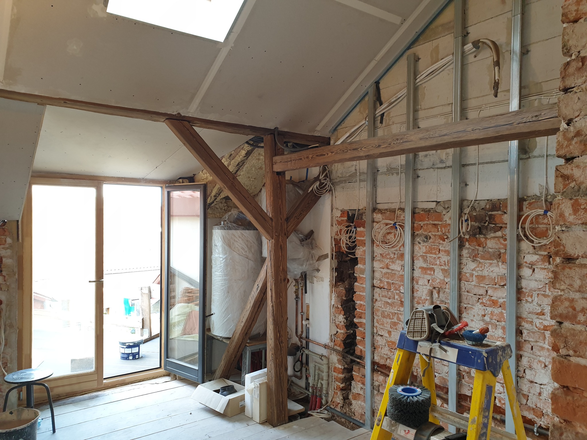 Wnętrze poddasza w trakcie remontu: widoczne odsłonięte cegły, drewniana konstrukcja więźby dachowej, instalacja elektryczna, drabina z narzędziami i otwarte drzwi balkonowe.
