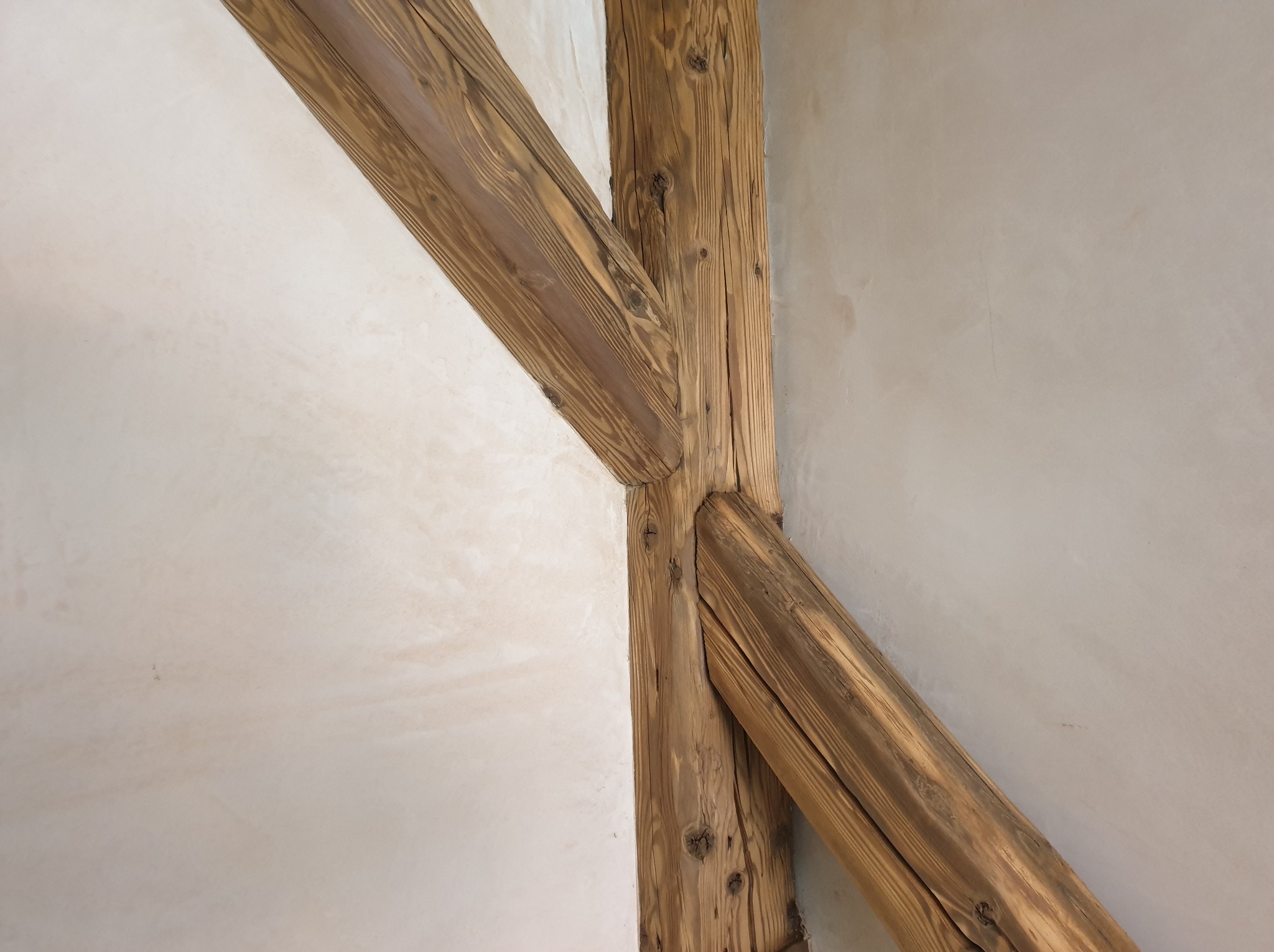 Drewniana konstrukcja wspierająca, łącząca się z ścianą o jasnym, kremowym wykończeniu, widoczne słoje drewna i naturalne niedoskonałości.