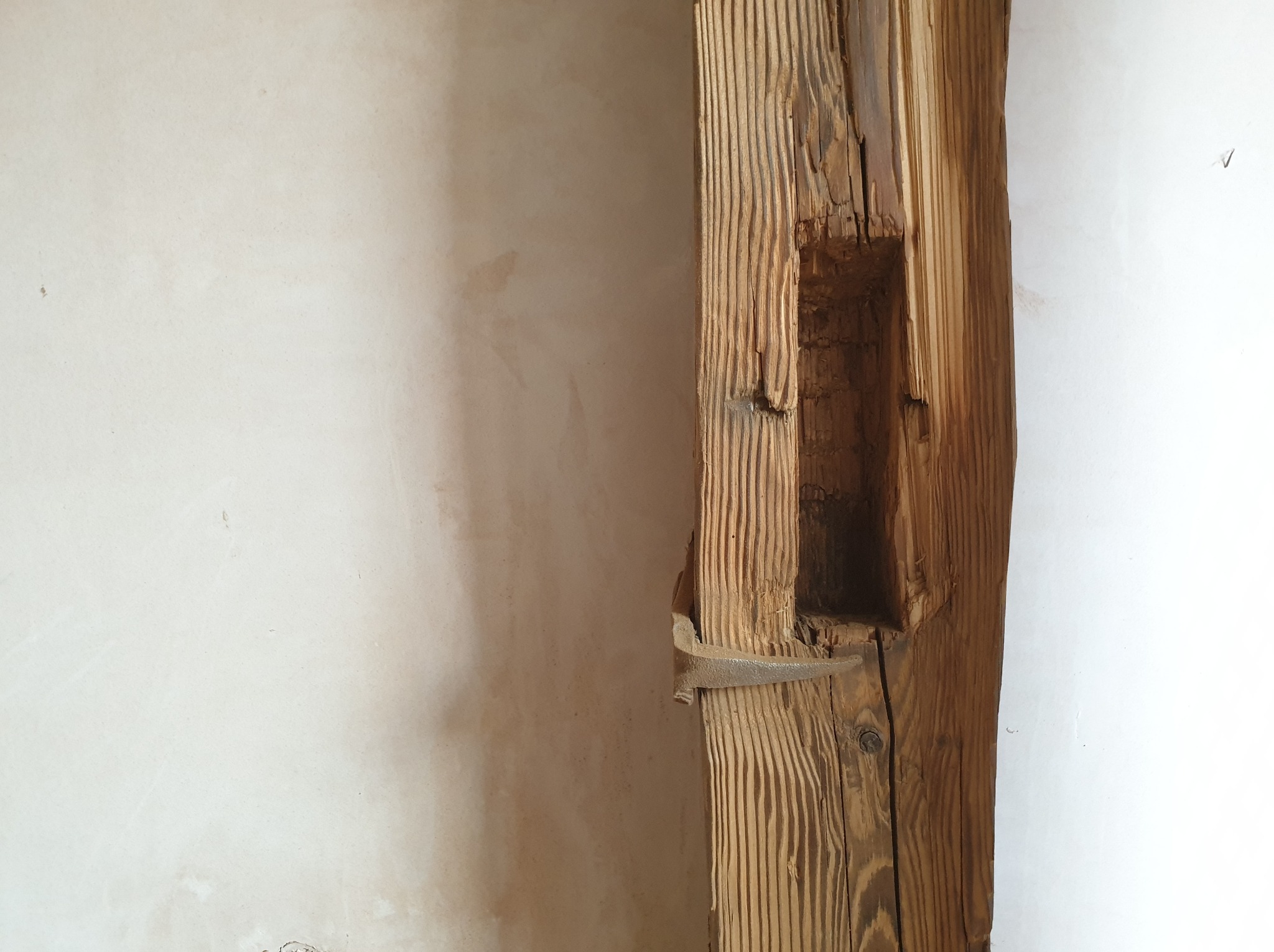 Fragment starej, drewnianej belki z widocznym wyżłobieniem i metalowym mocowaniem, przylegającej do pomalowanej ściany.