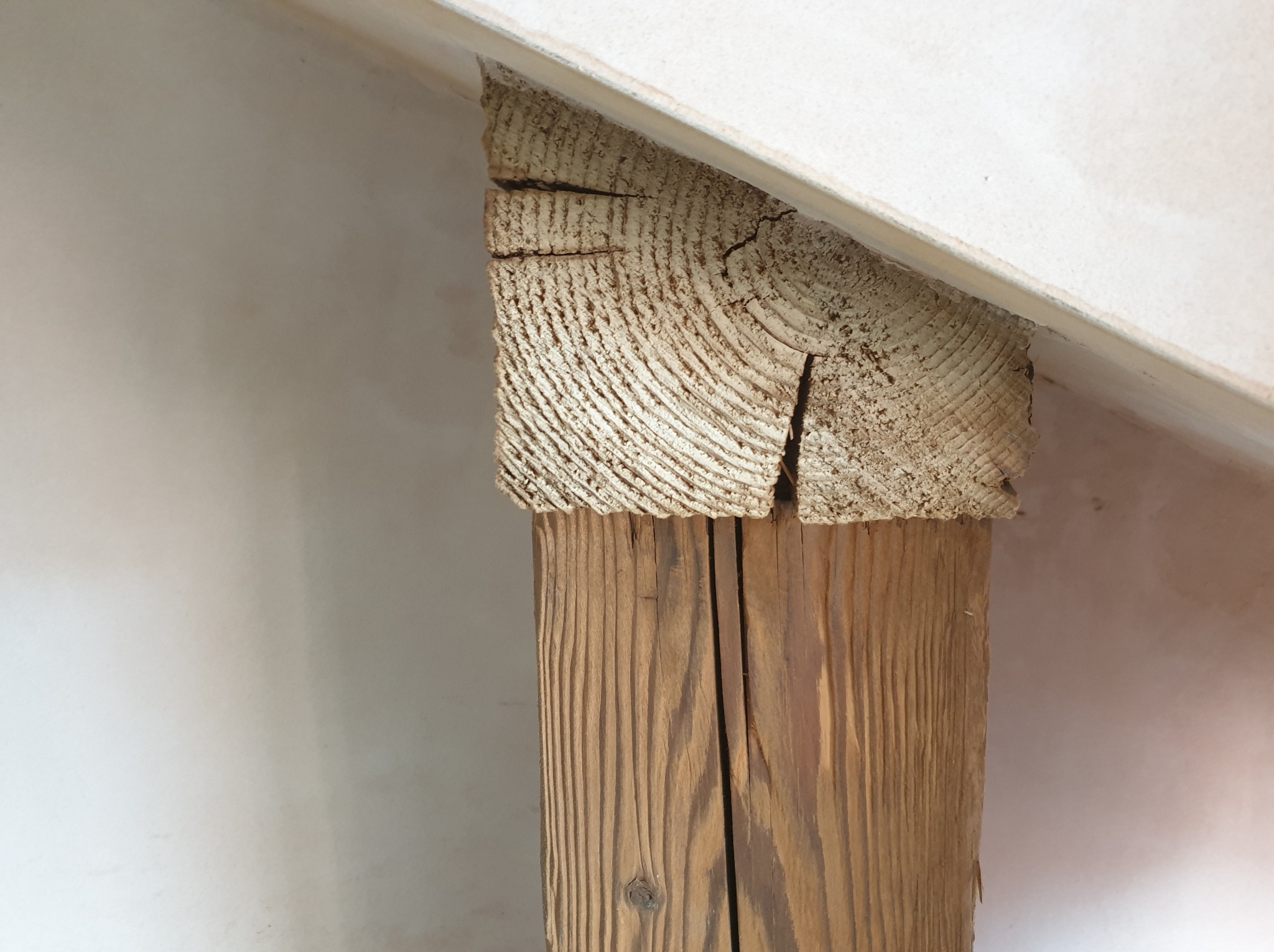 Drewniany słup konstrukcyjny z widocznymi słojami, podtrzymujący białą, ukośną powierzchnię sufitu, z widocznymi pęknięciami w drewnie.