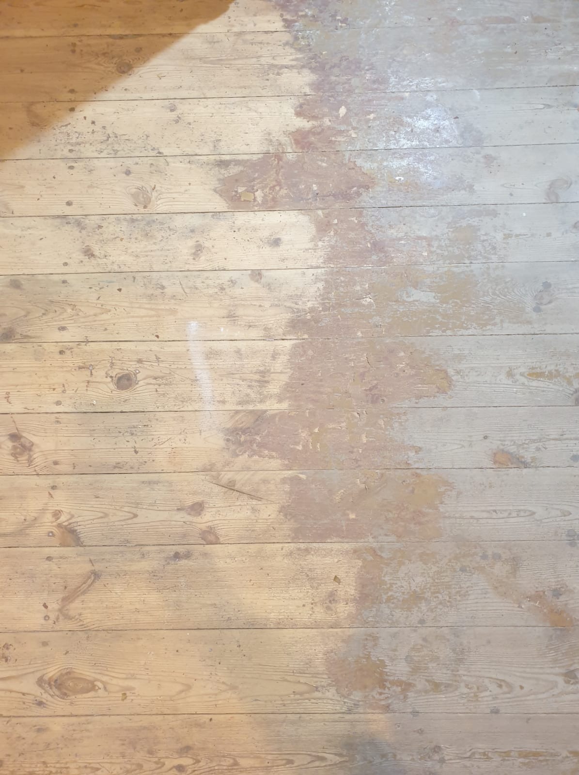 Stara, drewniana podłoga z widocznymi śladami uszkodzeń i ubytkami farby, ukazująca proces renowacji lub zużycie.