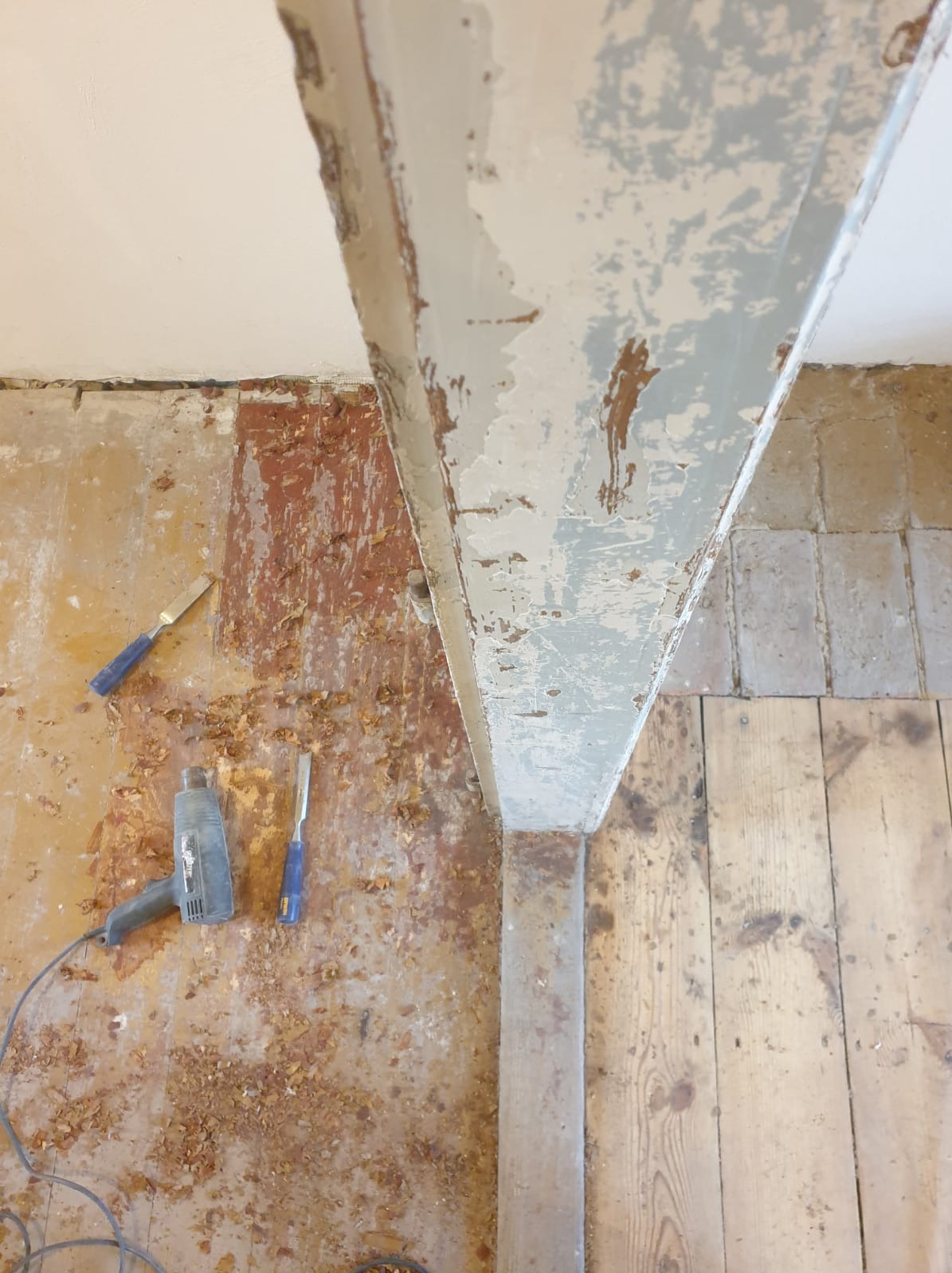 Fragment remontowanego pomieszczenia z drewnianą podłogą, ścianą z resztkami farby i odsłoniętą cegłą, oraz drewnianą belką w trakcie usuwania starej powłoki, widoczne narzędzia: opalarka i dłuta.