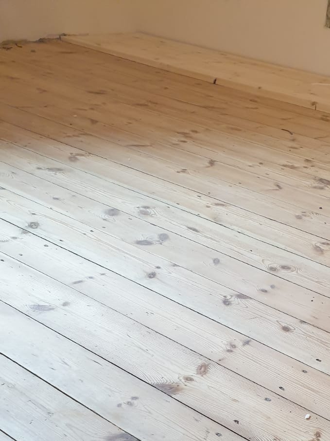 Jasne, surowe deski podłogowe z widocznymi sękami i śladami po szlifowaniu, ułożone równolegle do ściany w tle.