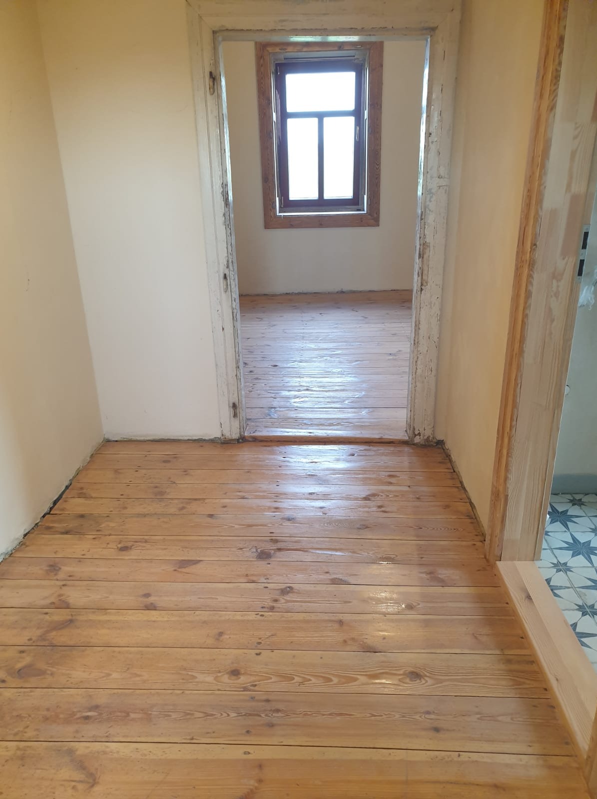 Odnowiona drewniana podłoga z widocznymi słojami, świeżo polakierowana, z widokiem na okno w następnym pomieszczeniu przez odrestaurowane, białe futryny drzwi.