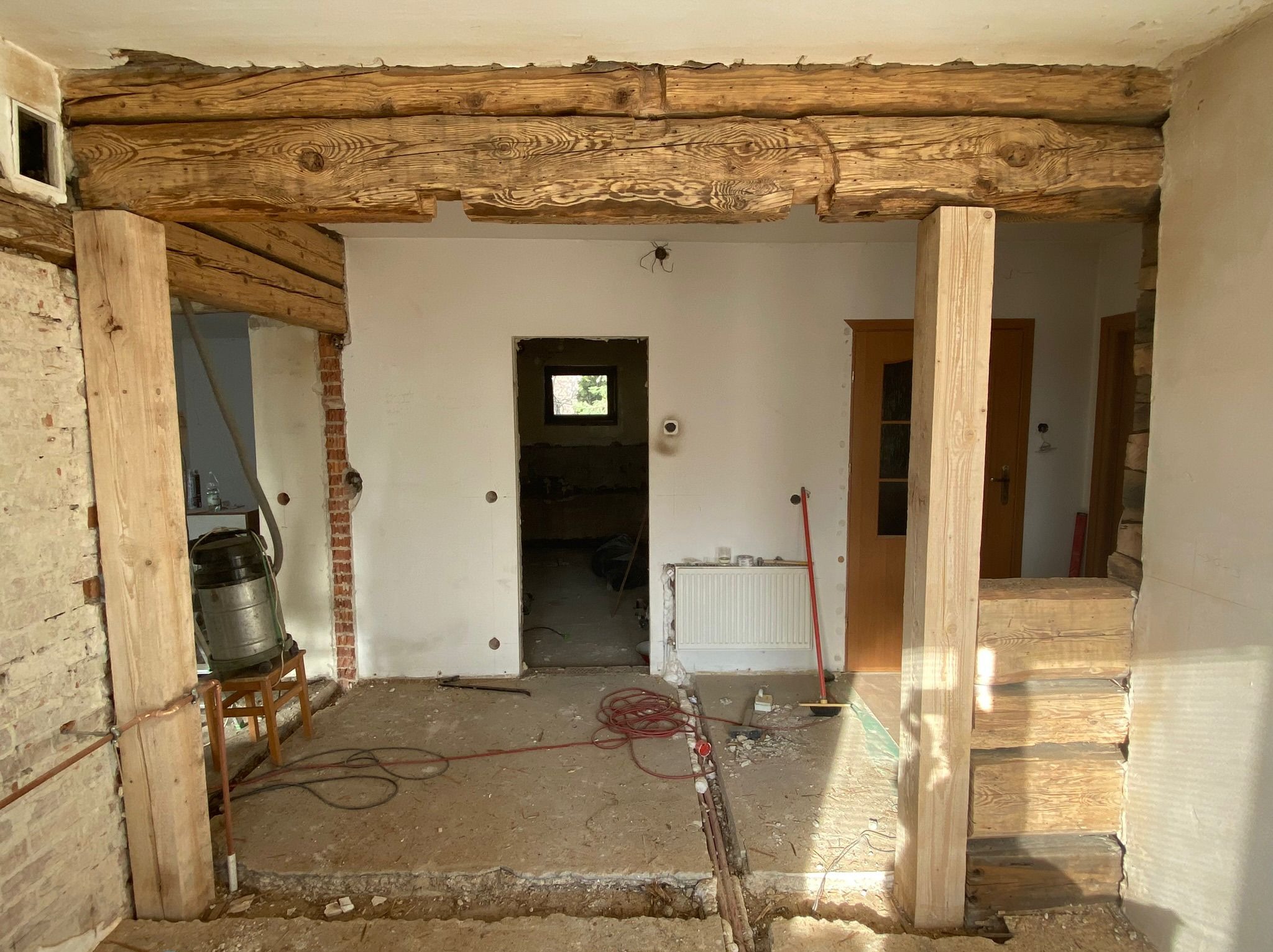 Wnętrze w trakcie remontu z odsłoniętymi drewnianymi belkami stropowymi, wspartymi na pionowych słupach. Widoczne instalacje w podłodze, odsłonięta cegła i przygotowania do dalszych prac...