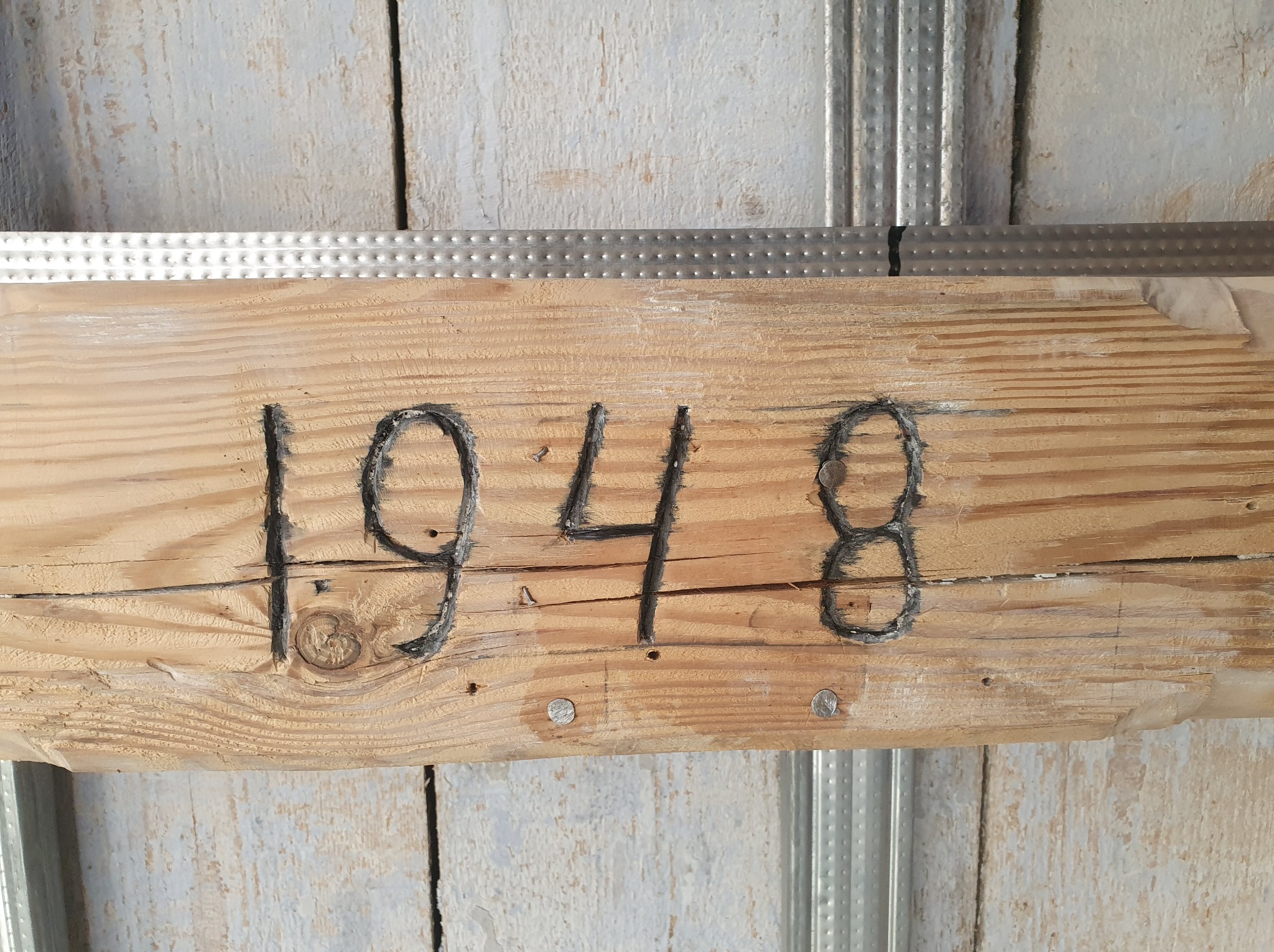 Surowe drewno z wypalonym numerem '1948' przymocowane do metalowej konstrukcji sufitowej, widoczne ślady obróbki i niedoskonałości drewna.