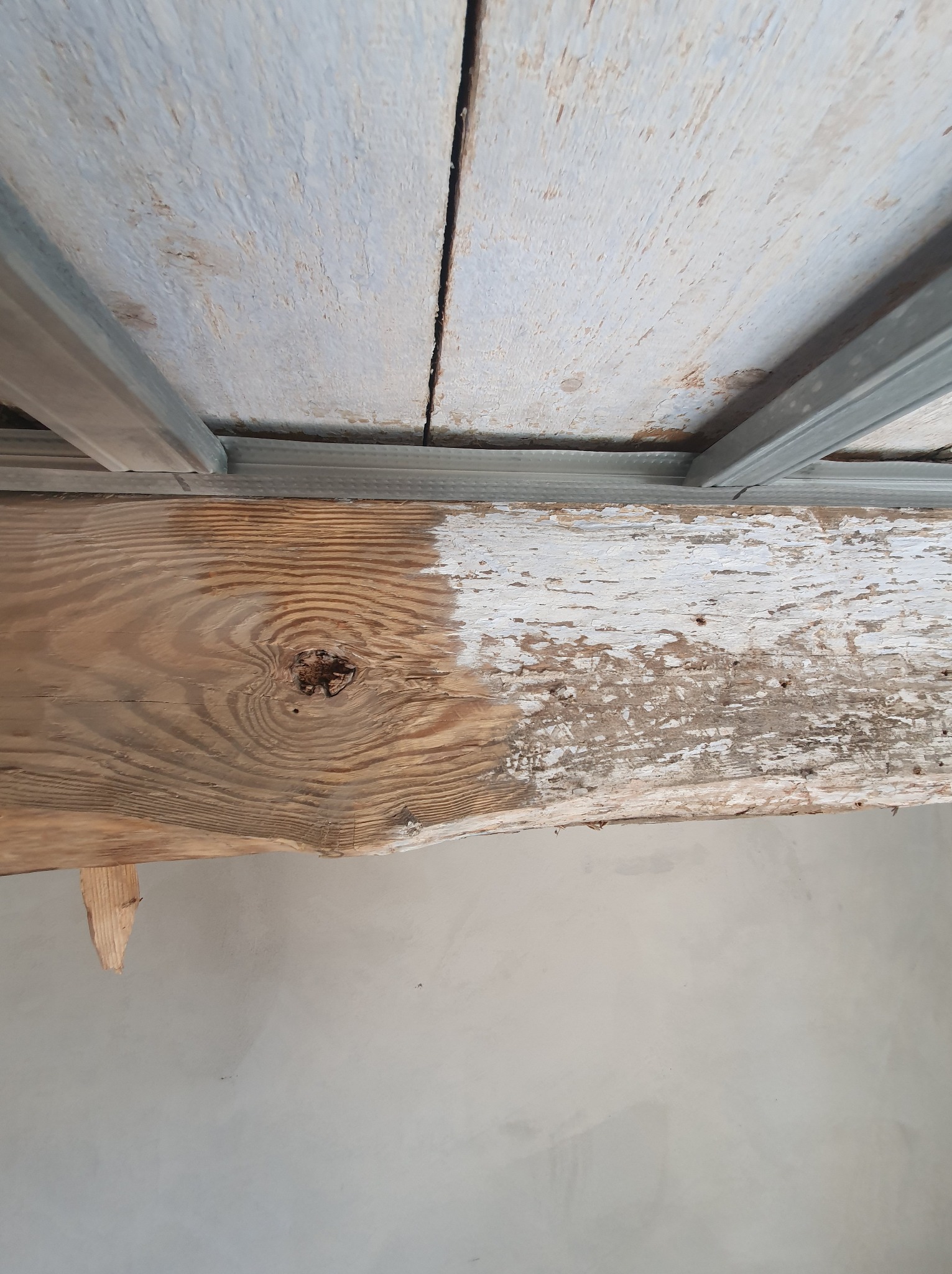 Surowe drewno więźby dachowej z widocznym przekrojem i fragmentami starej, łuszczącej się farby, wsparte na metalowym profilu, z widocznym fragmentem sufitu i ściany w tle.