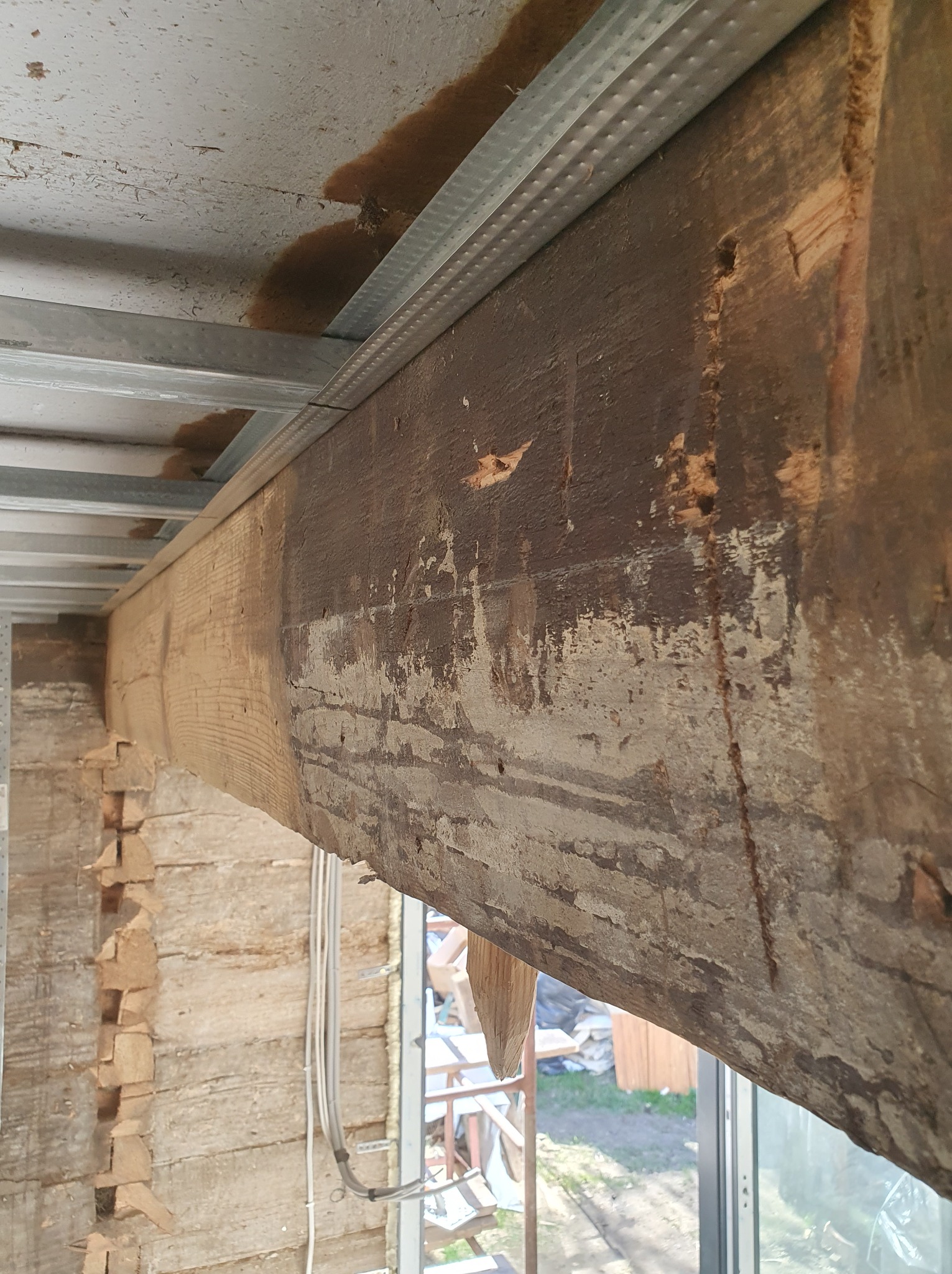 Drewniana belka stropowa z widocznymi śladami obróbki i częściowo zachowaną starą powłoką, podtrzymywana przez metalową konstrukcję sufitu podwieszanego, w tle fragment ściany z bali.