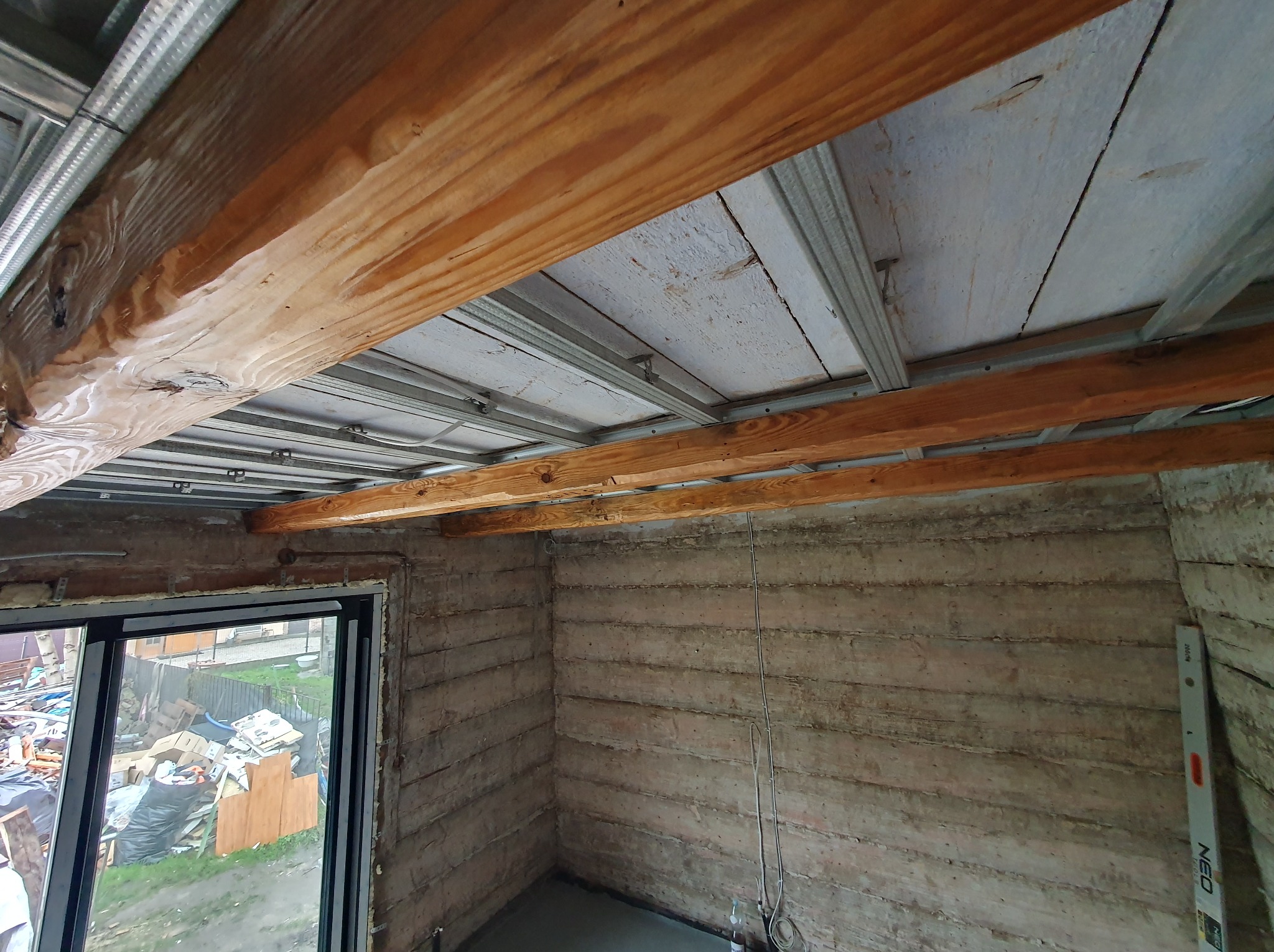 Wnętrze w trakcie remontu: surowe ściany z poziomnicą, drewniane belki stropowe podwieszone na metalowej konstrukcji, widok na zewnątrz przez okno z gruzem na trawniku.