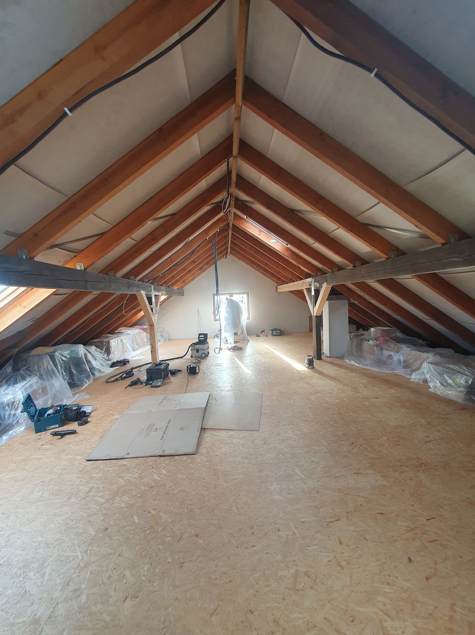 Poddasze w trakcie adaptacji z widoczną konstrukcją więźby dachowej, ociepleniem, nową podłogą z płyt OSB oraz narzędziami przygotowanymi do dalszych prac wykończeniowych.
