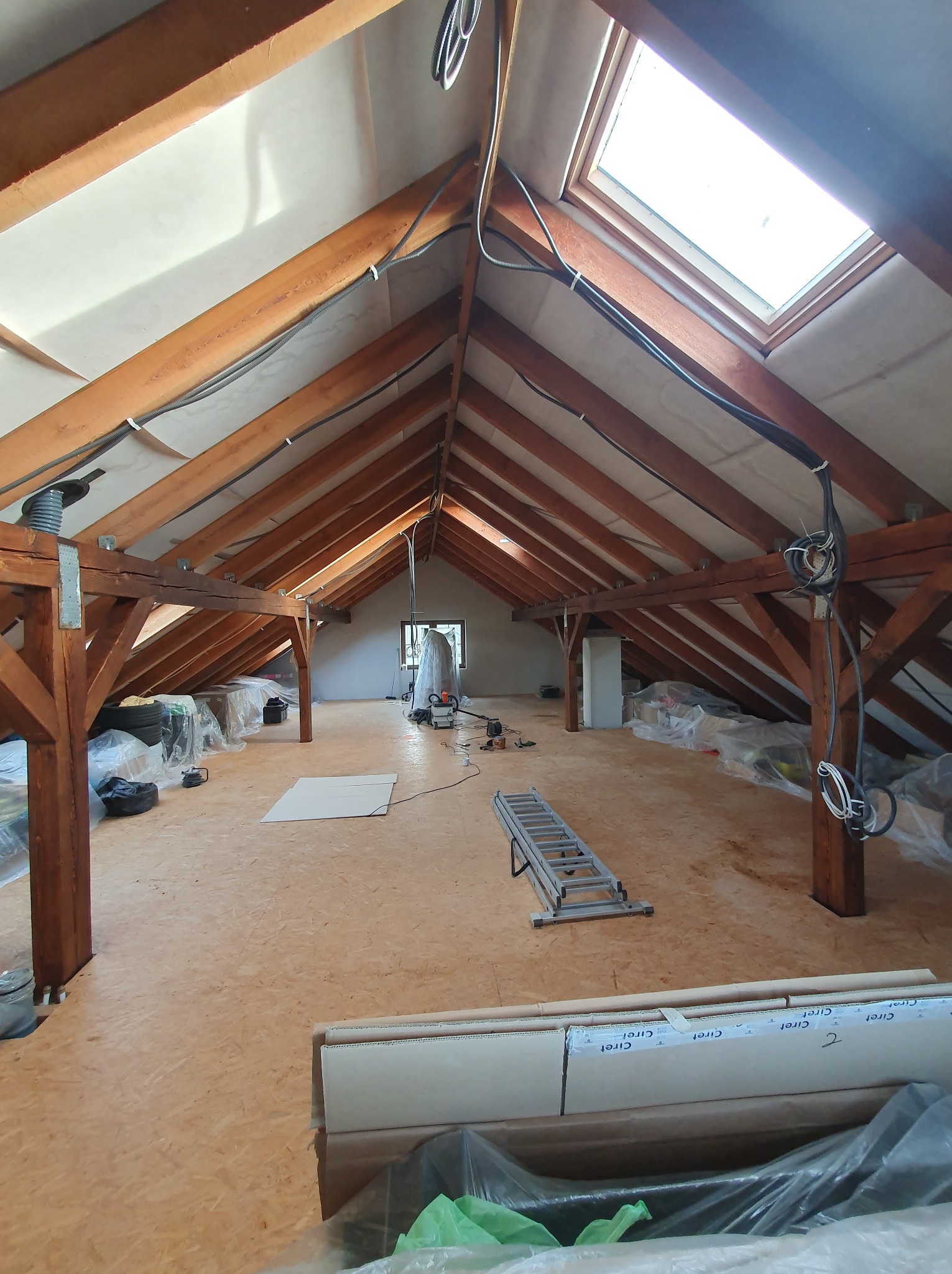 Drewniana konstrukcja dachu poddasza z oknem dachowym, widoczne belki, izolacja, kable elektryczne oraz przygotowane materiały budowlane i drabina.