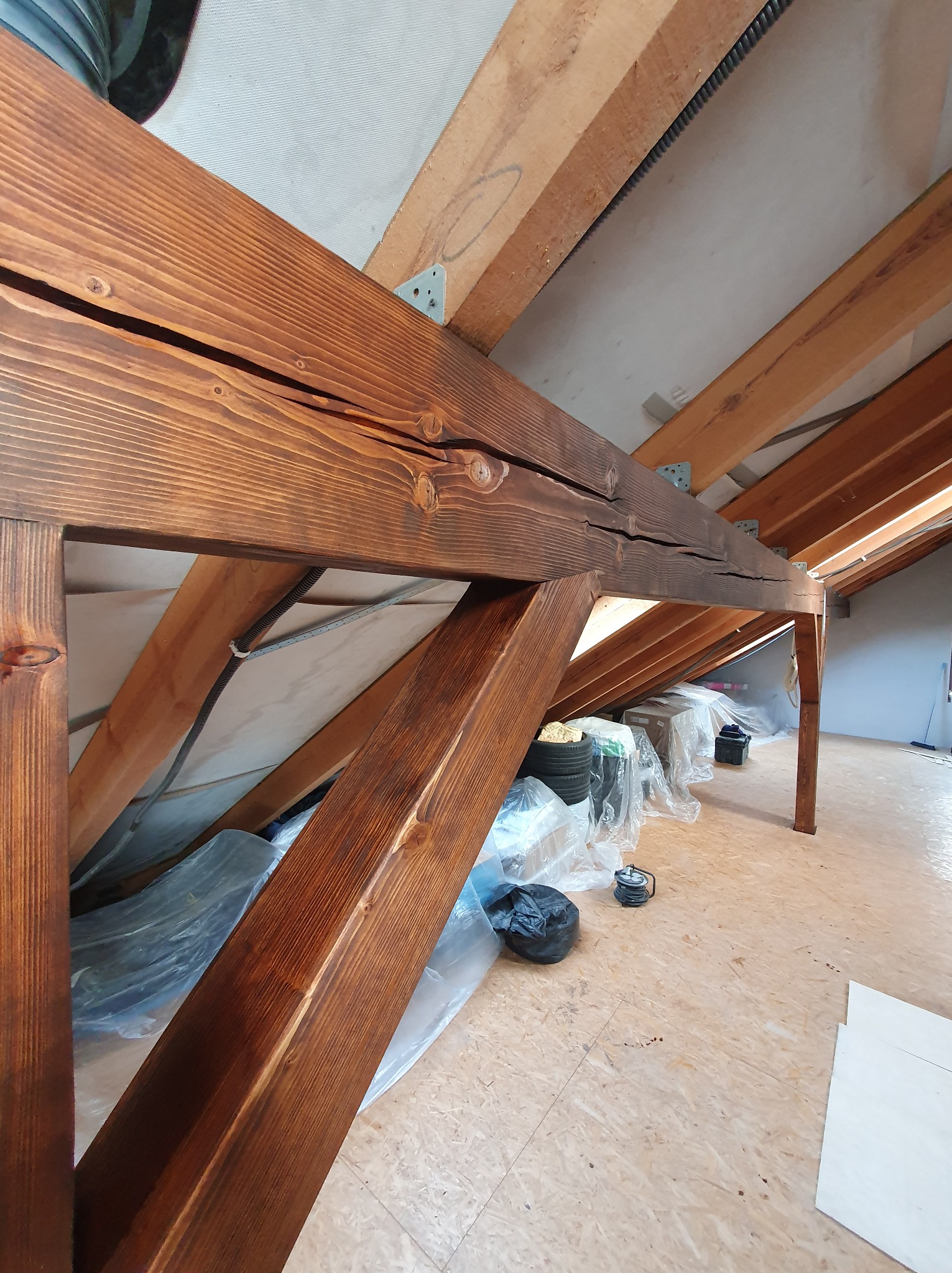 Widok na konstrukcję więźby dachowej z belek zaimpregnowanych na brązowo, widoczne metalowe łączniki, część poddasza zabezpieczona folią.