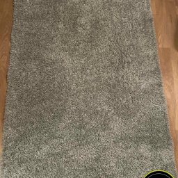 Fresh Clean - Szary dywan typu shaggy leżący na drewnianej podłodze, widoczne logo firmy czyszczącej w prawym dolnym rogu.