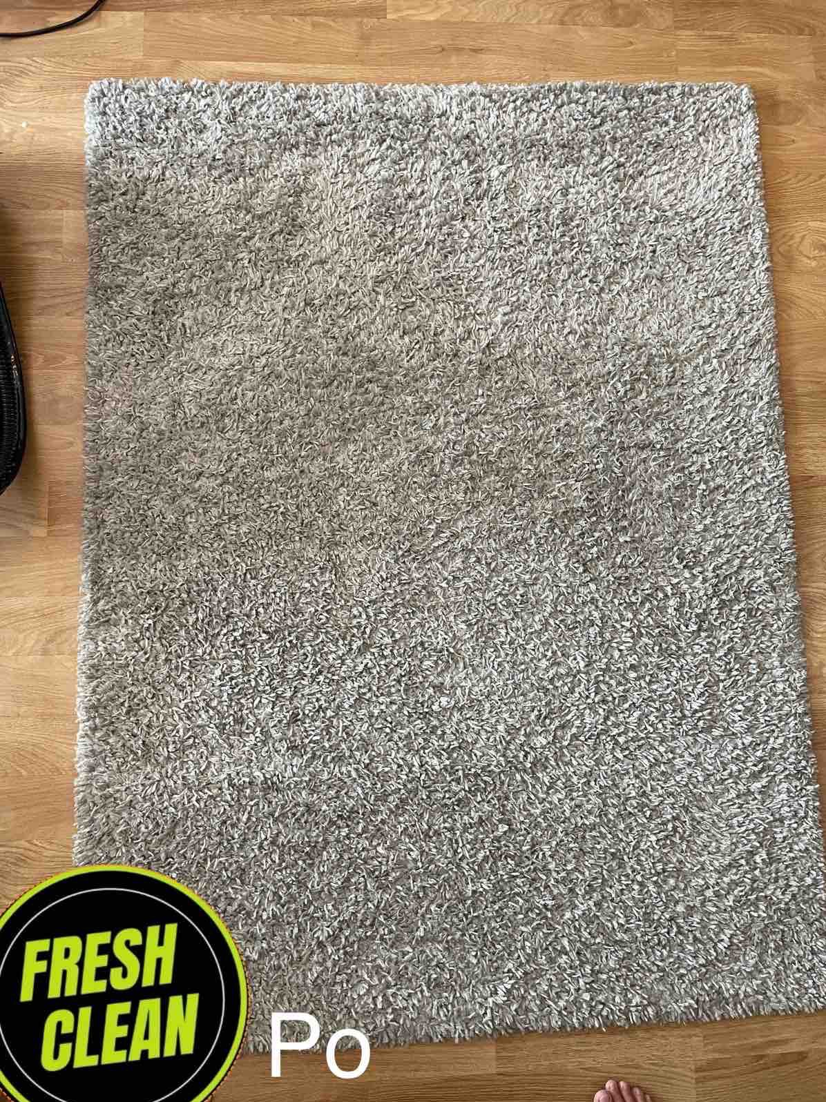 Wyprany, puszysty, szary dywan typu shaggy leżący na drewnianej podłodze, z widocznym napisem 'FRESH CLEAN Po' w rogu.