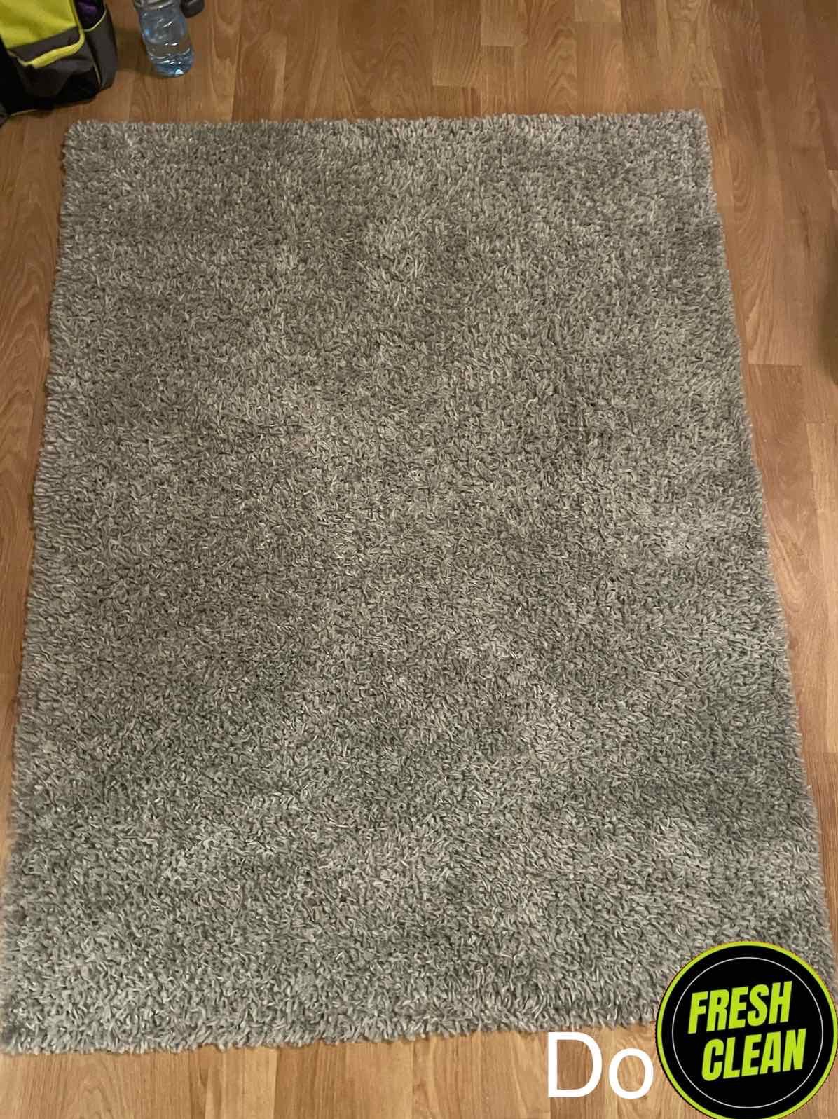 Szary dywan typu shaggy leżący na drewnianej podłodze, widoczne logo firmy czyszczącej w prawym dolnym rogu.
