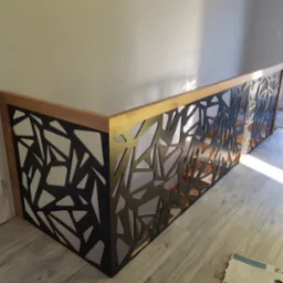 Nowoczesna balustrada z czarnego metalu o geometrycznym wzorze, osadzona na drewnianej ramie, oświetlona naturalnym światłem w jasnym wnętrzu.