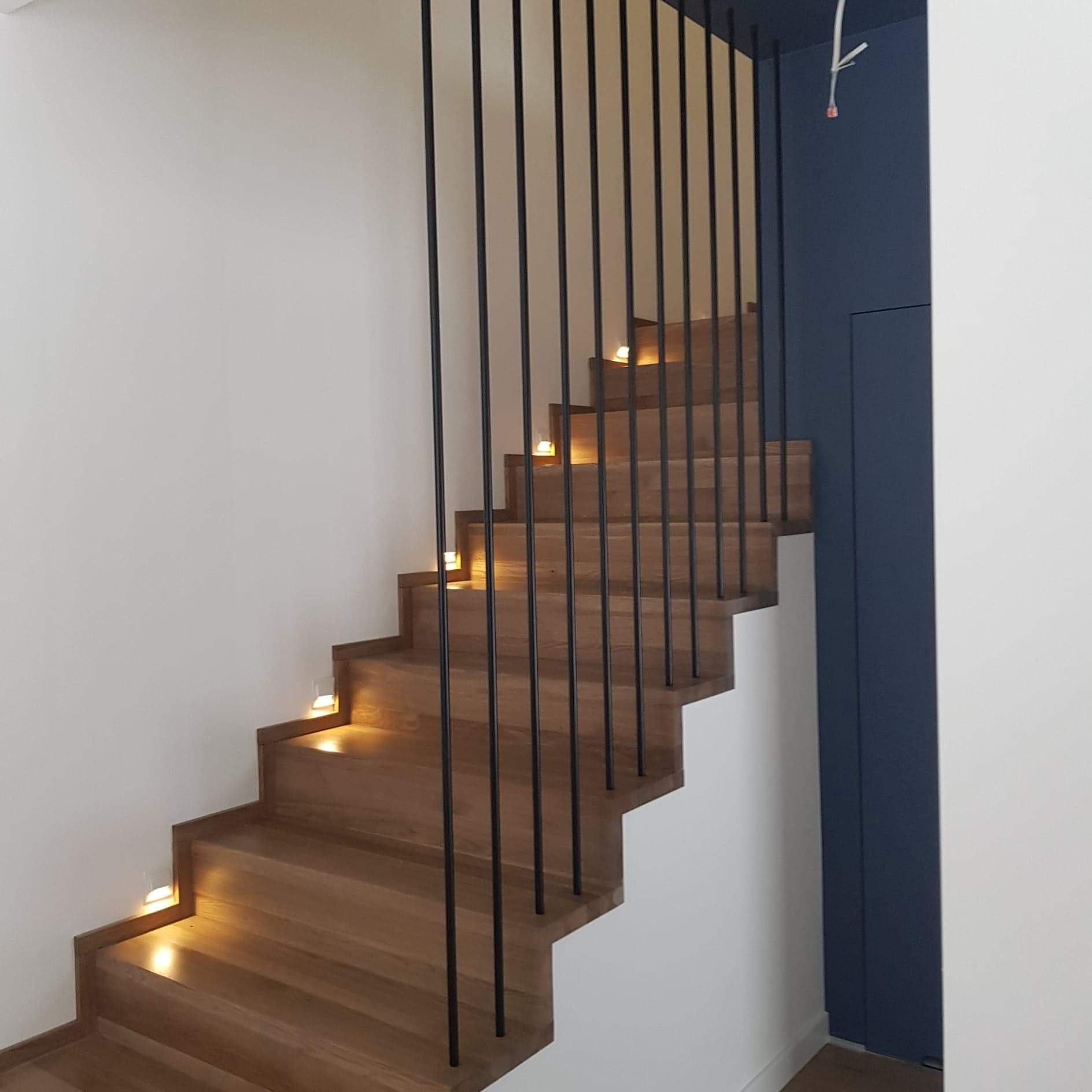 Nowoczesne schody z drewna z metalowymi balustradami, podświetlane stopnie, minimalistyczny design, białe i granatowe ściany.