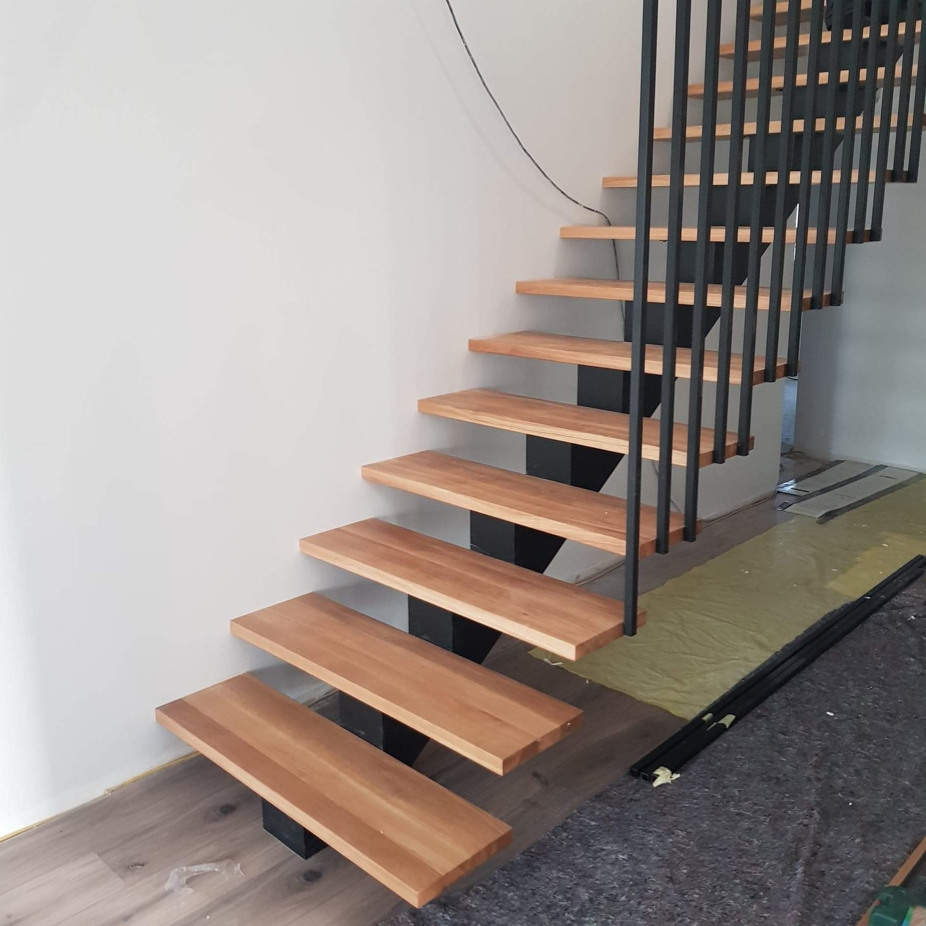 Nowoczesne schody z drewnianymi stopniami i czarną metalową balustradą, widoczne elementy montażowe i zabezpieczenia na podłodze.