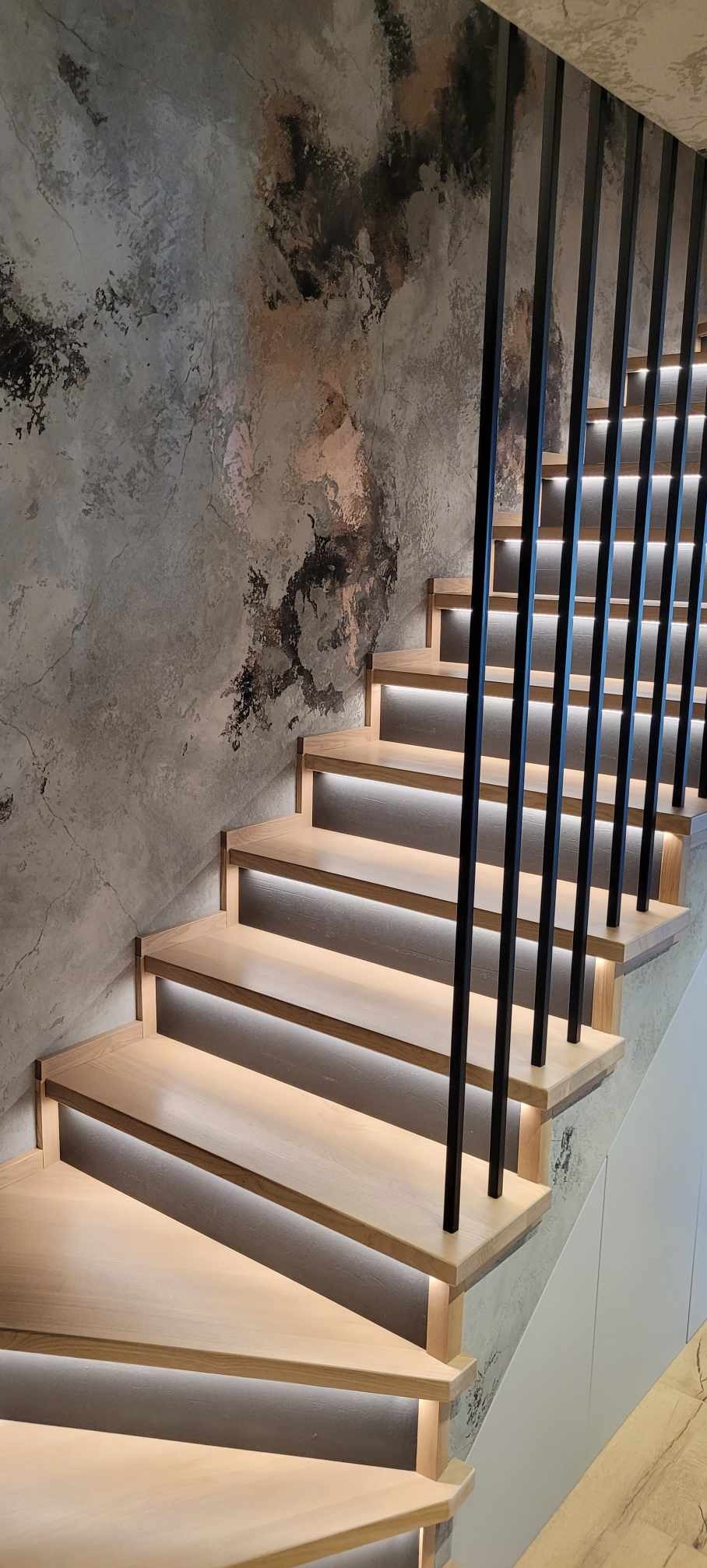 Nowoczesne schody z jasnego drewna z podświetleniem LED pod każdym stopniem i czarną, minimalistyczną balustradą na tle ściany o fakturze betonu.