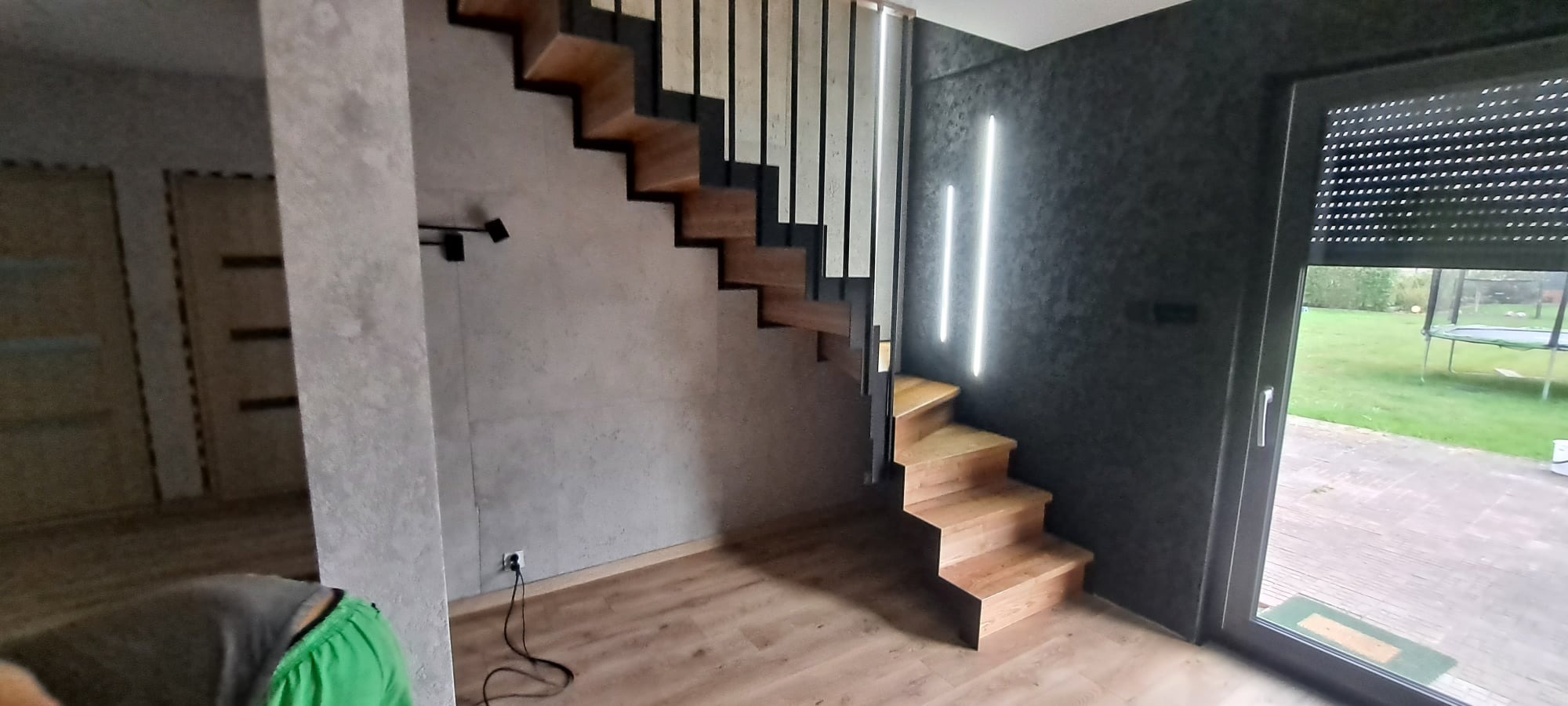Nowoczesne schody z drewnianymi stopniami i metalową balustradą, oświetlone pionowymi lampami LED na ciemnej ścianie, widok na ogród przez okno.