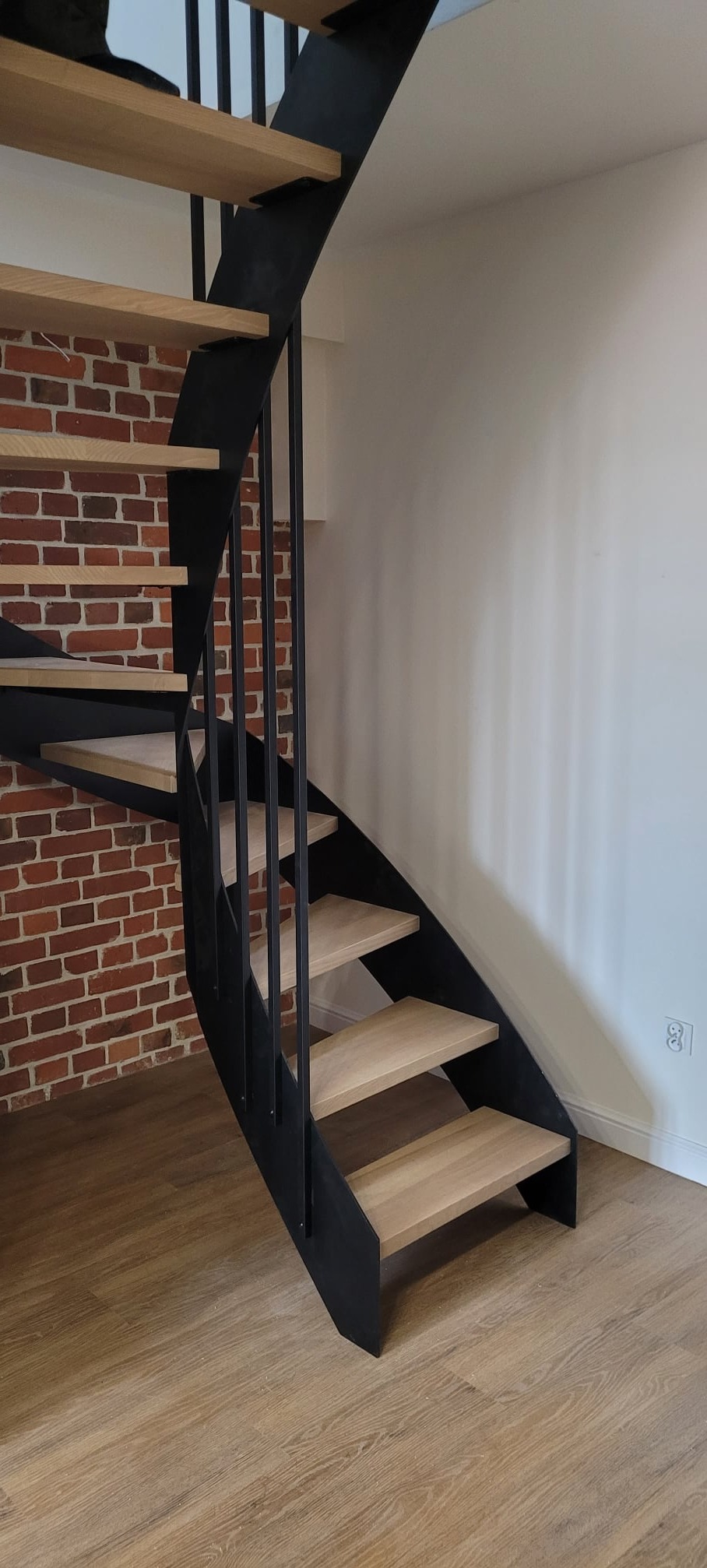 Spiralne schody z drewnianymi stopniami i czarną metalową konstrukcją, widoczne na tle ceglanej ściany i białej ściany z gniazdkiem elektrycznym.
