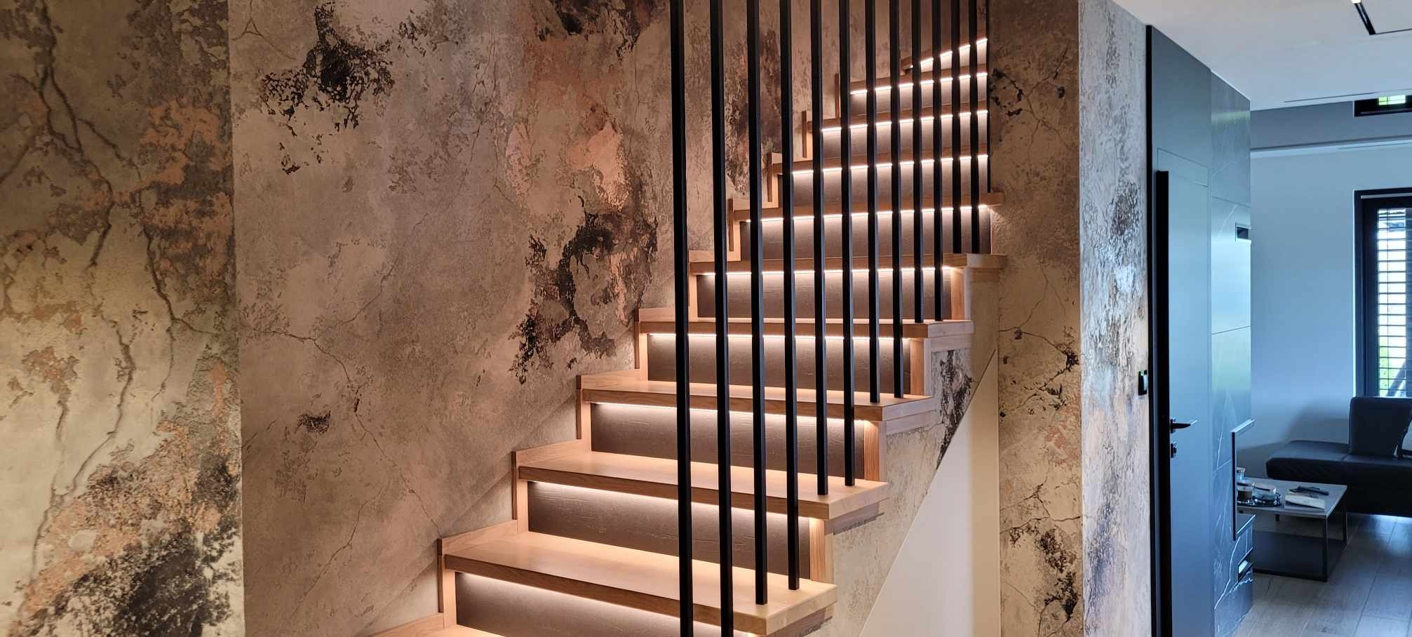 Nowoczesne schody z podświetleniem LED stopni i czarnymi metalowymi balustradami, na tle ściany z efektem betonu.